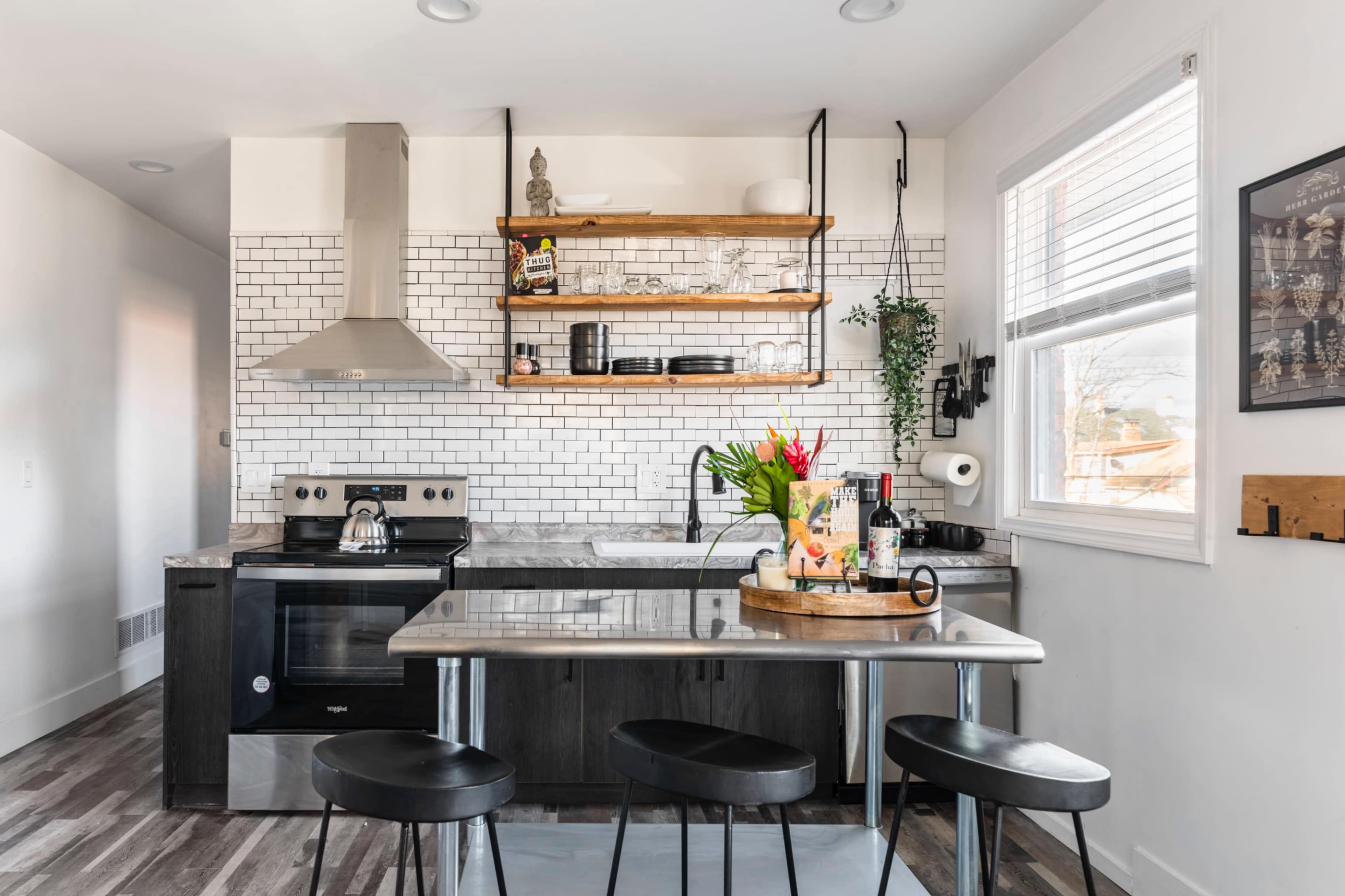 A modern kitchen features a stainless steel stove, open wooden shelving with dishes, a gray marble countertop, and a round table with a floral centerpiece.