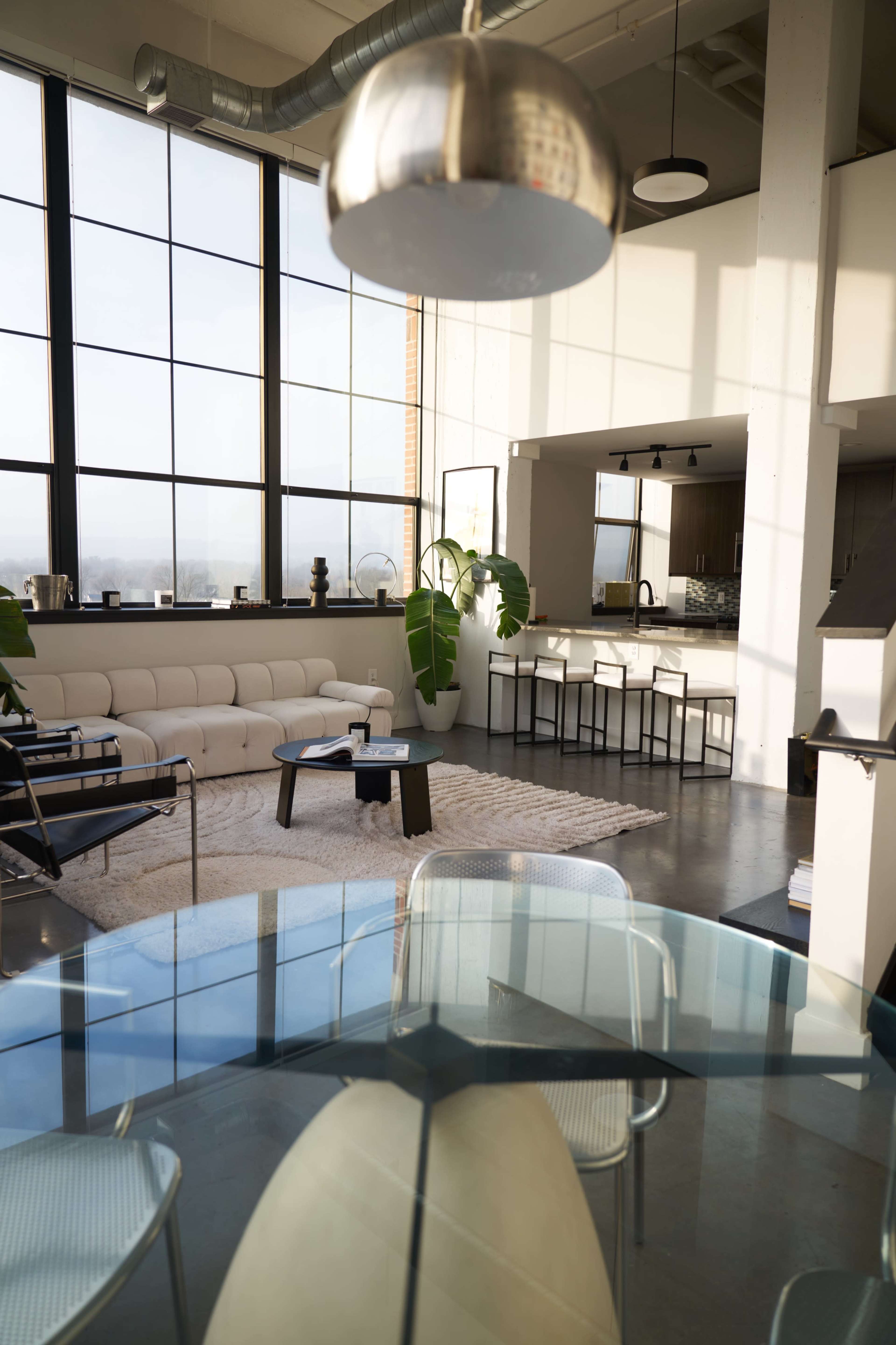 A modern open-concept living space features large windows, a sofa, a dining area, and a kitchen with a bar counter.