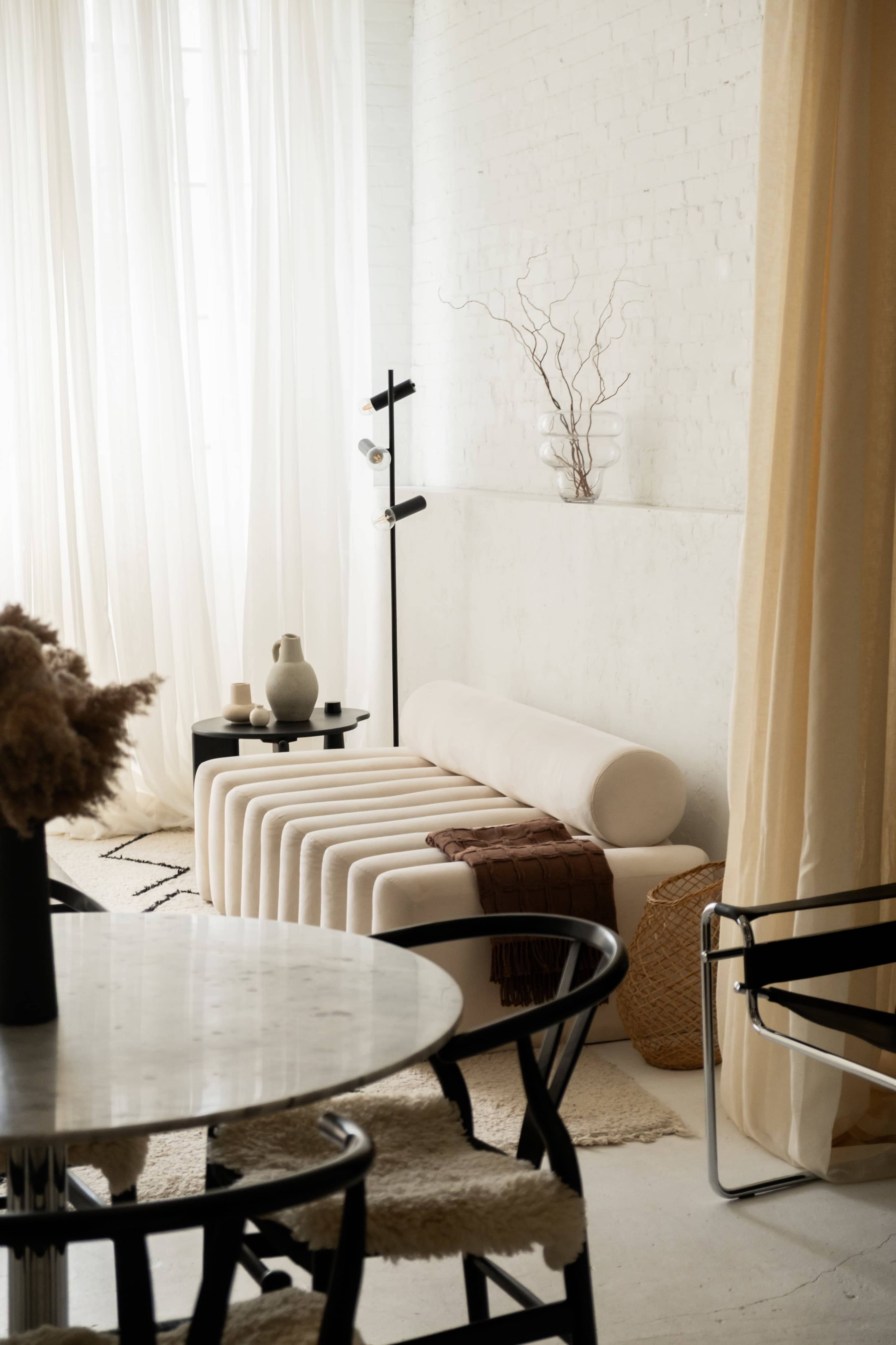 The image shows a modern interior with a light-colored, sculptural sofa, a round dining table with black chairs, and soft drapes allowing natural light to enter the space.