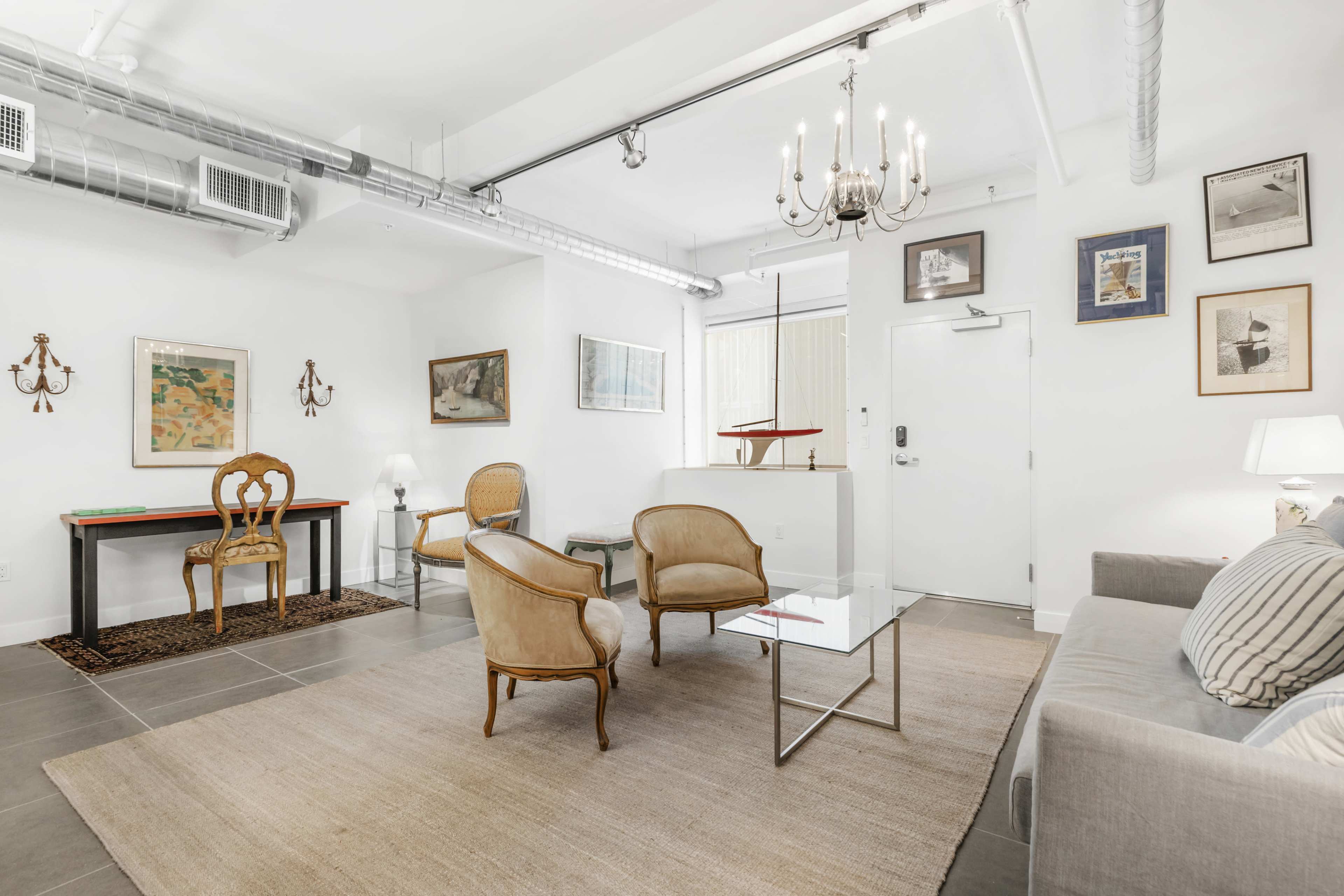 The image shows a modern living room with a sofa, two chairs, a glass coffee table, and various framed artworks on the white walls.