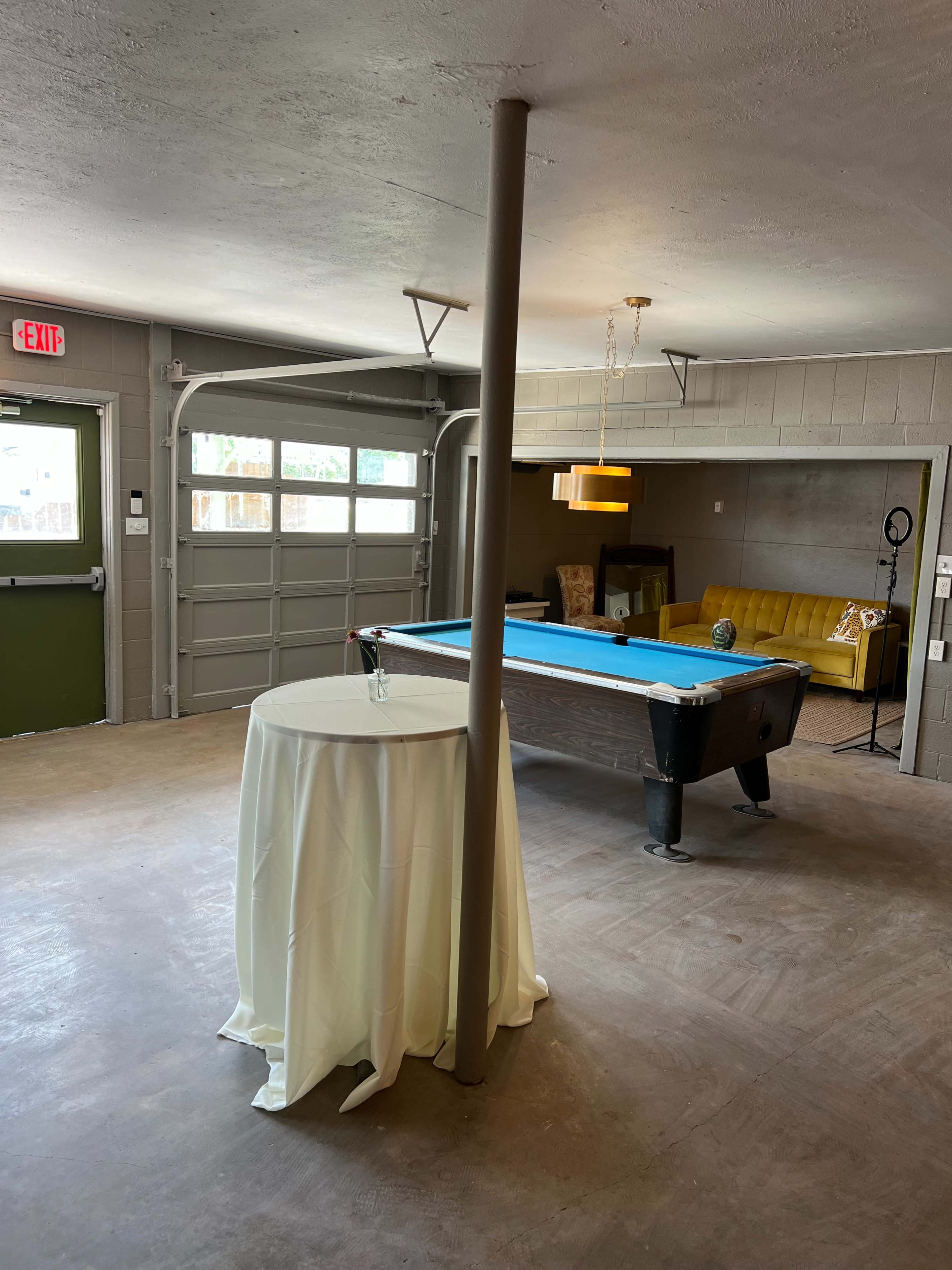 The image shows a garage converted into a recreation room, featuring a pool table, a yellow couch, a round table with a white tablecloth, and a hanging light fixture.