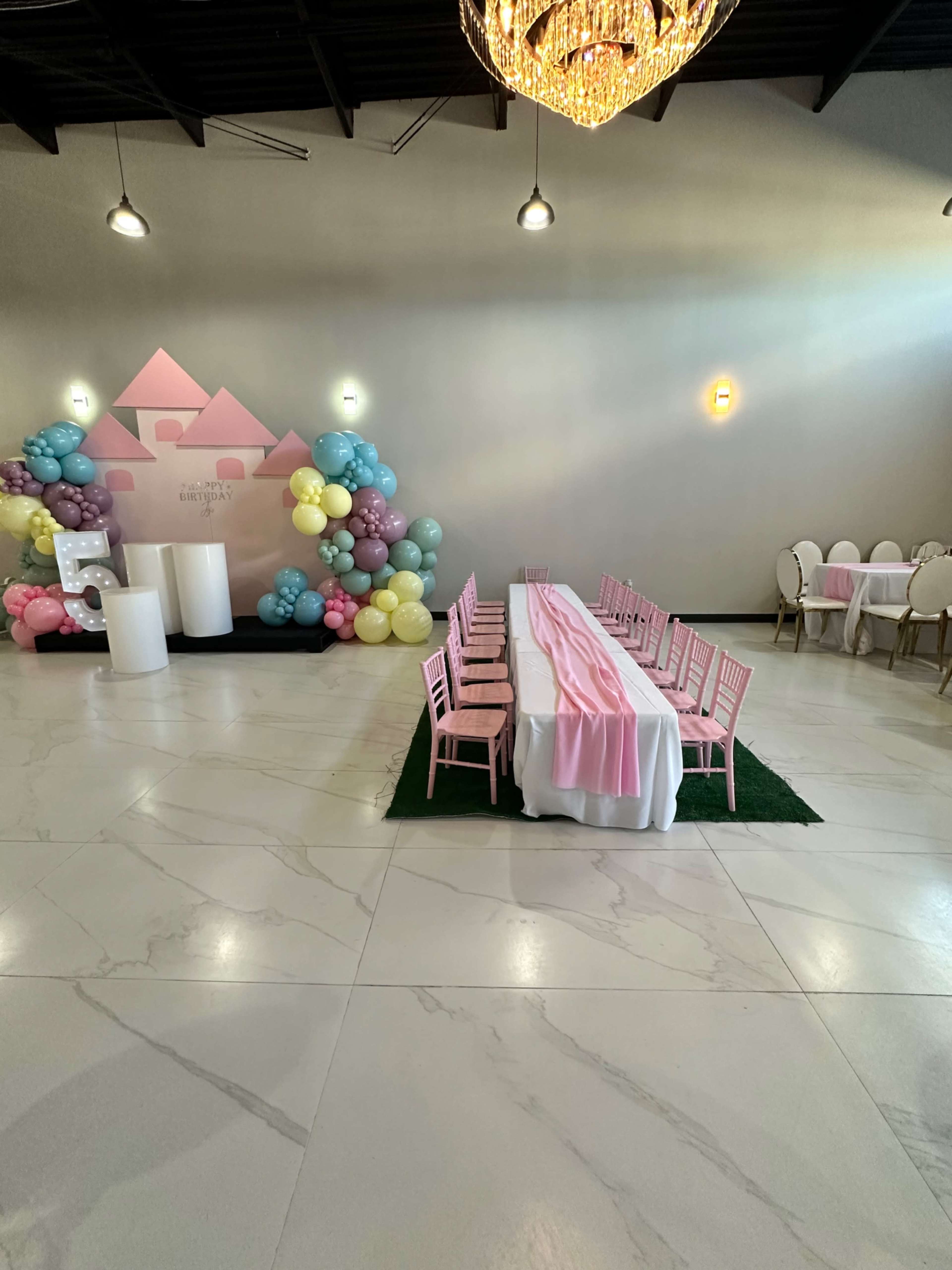 A decorated indoor event space featuring a long table set with pink tablecloths, surrounded by pink chairs, and balloon decorations in pastel colors at one end.