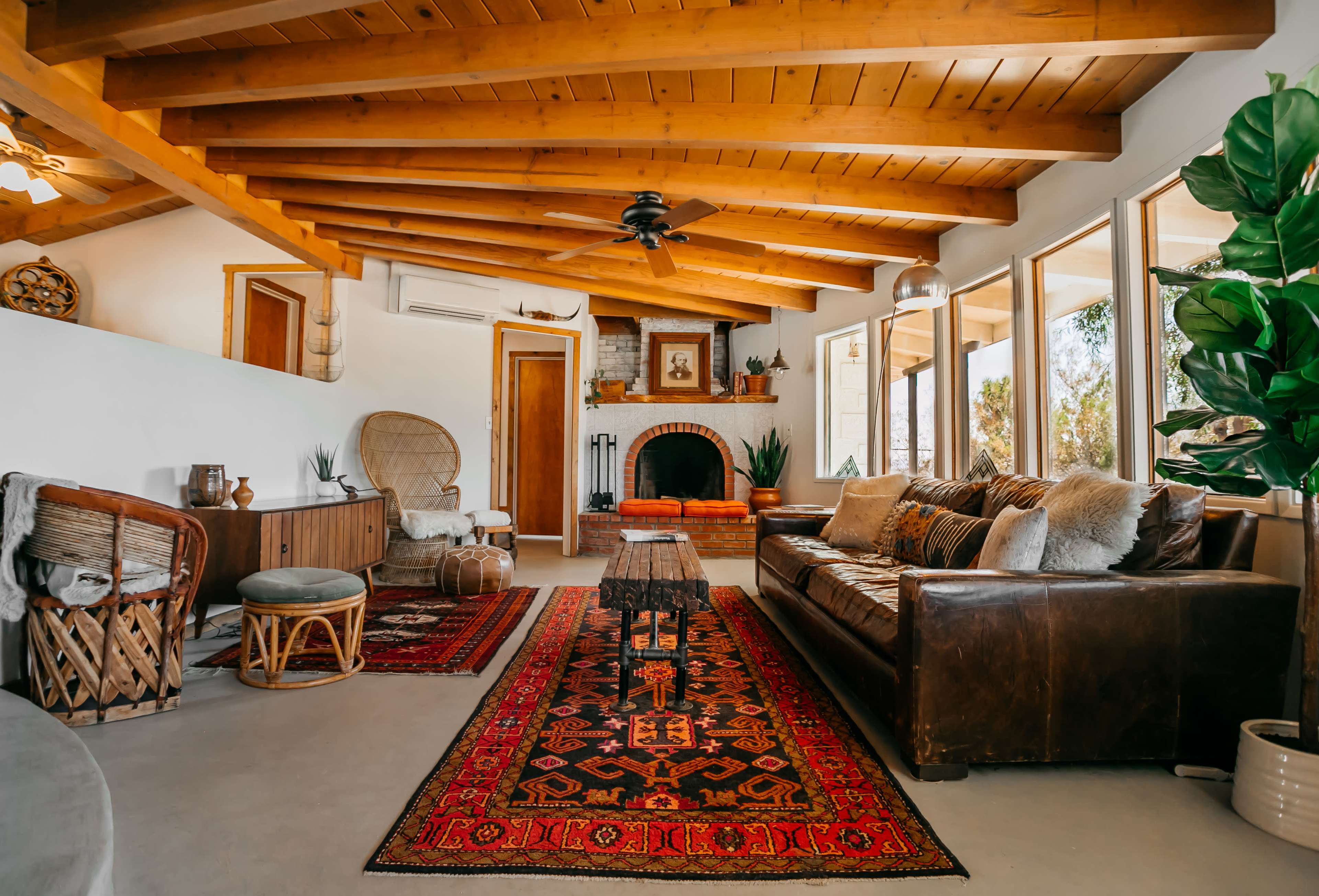 A cozy living room features a leather sofa, an area rug, wooden beams on the ceiling, and large windows with views of greenery.