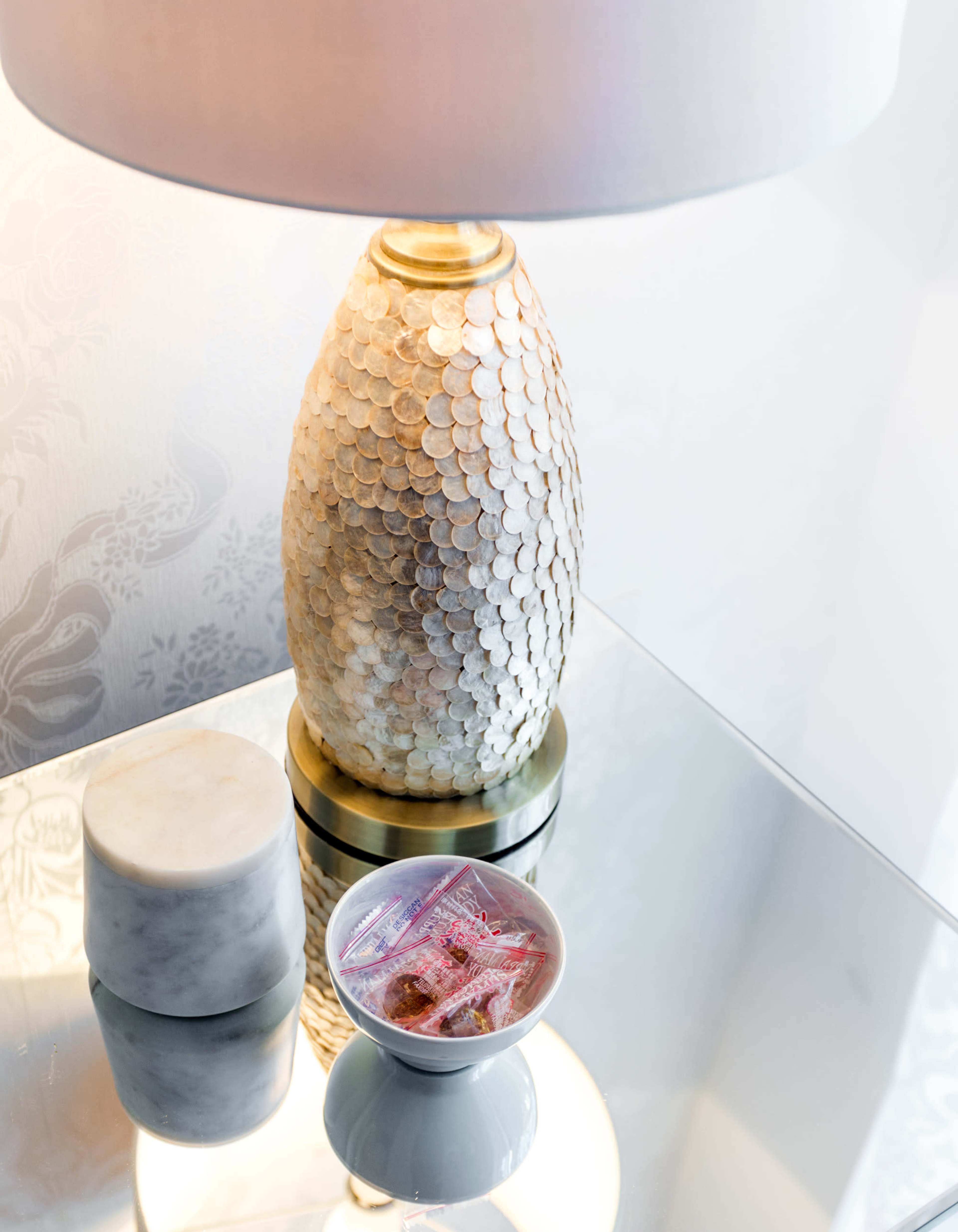 A lamp with a decorative base made of circular shells sits next to a marble container and a small dish holding colorful candies on a reflective surface.