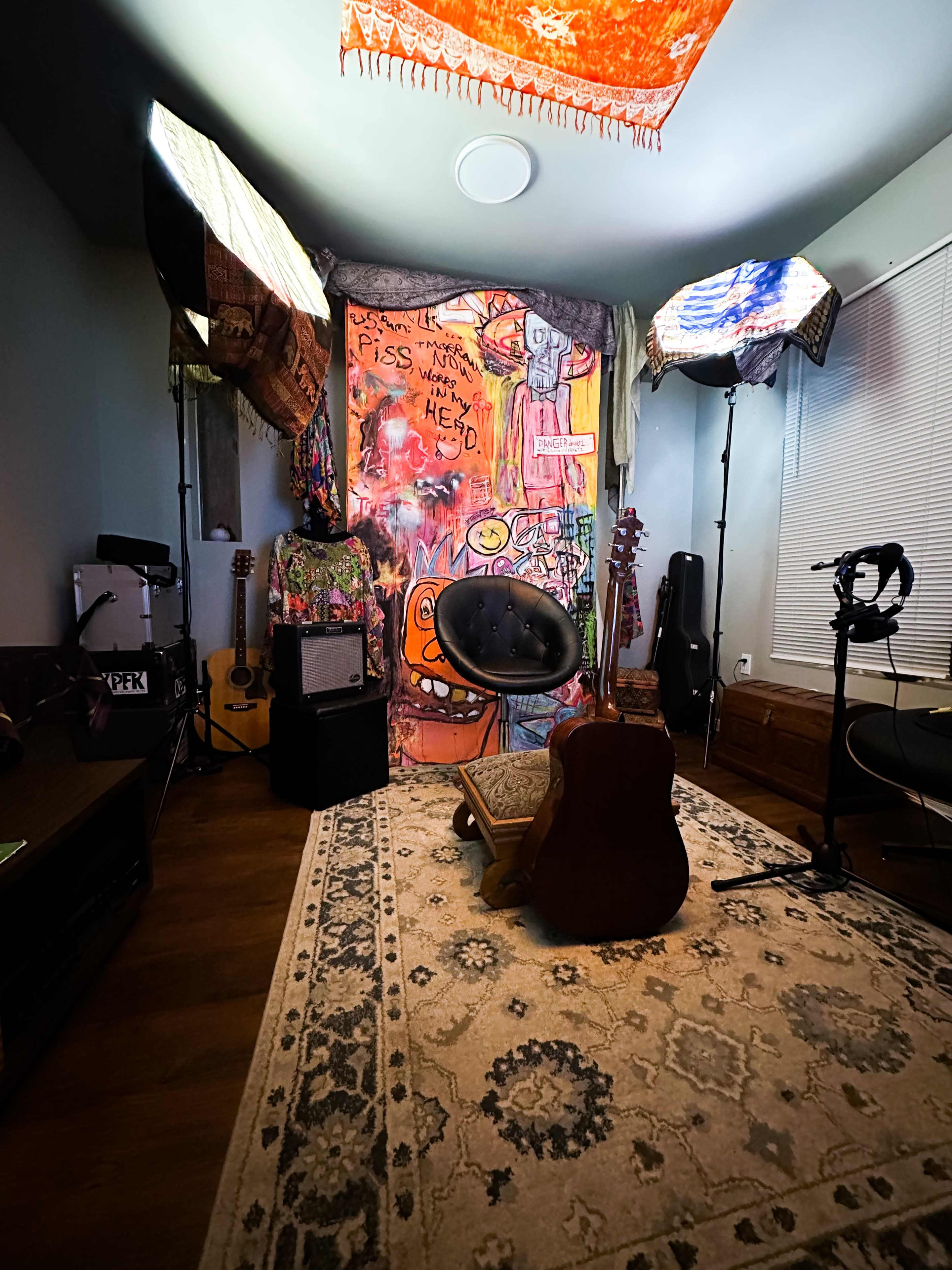The image depicts a music studio or practice space featuring a black chair in front of a brightly decorated backdrop, guitars leaning against walls, and lighting equipment set up.