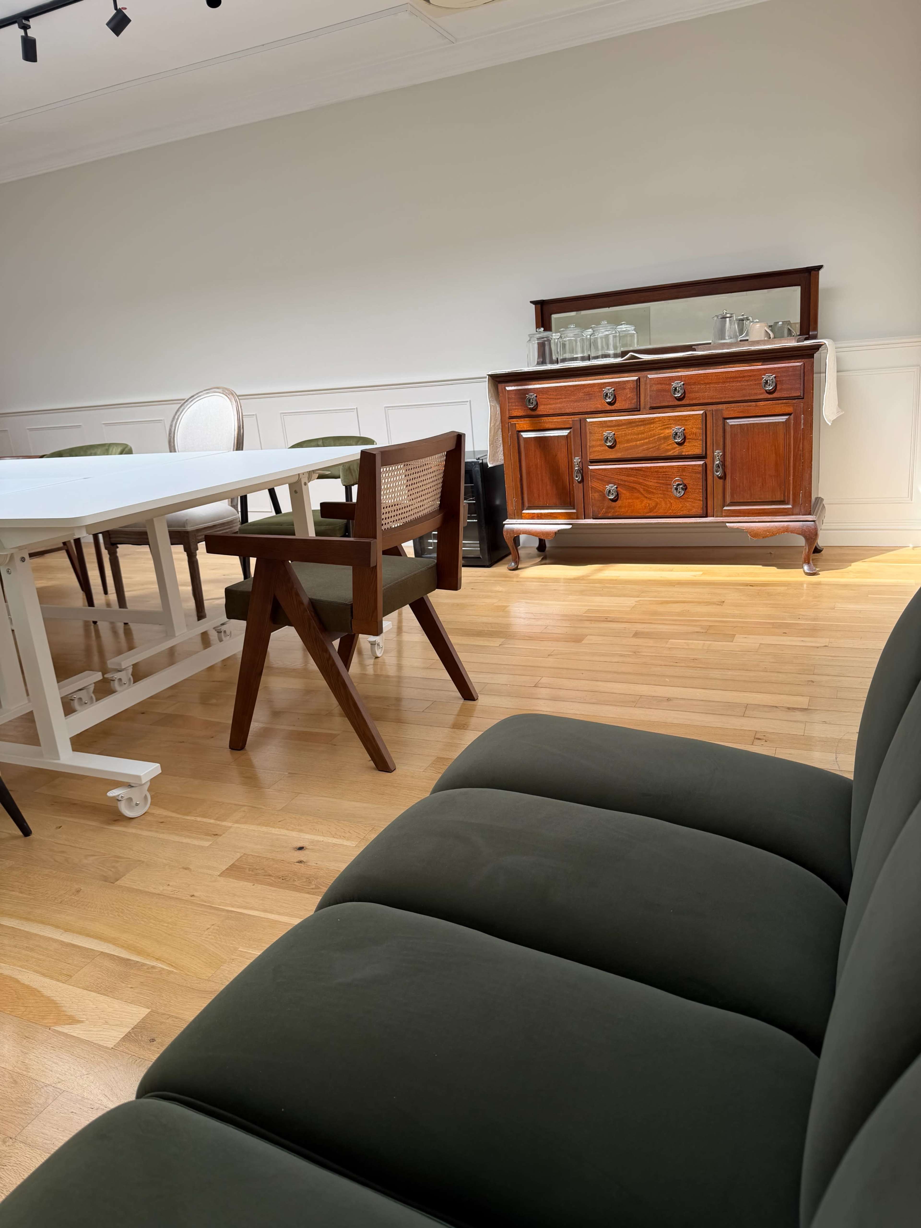 The image shows a spacious room with wooden flooring, a wooden sideboard against the wall, and a dining area with various chairs and a white table.