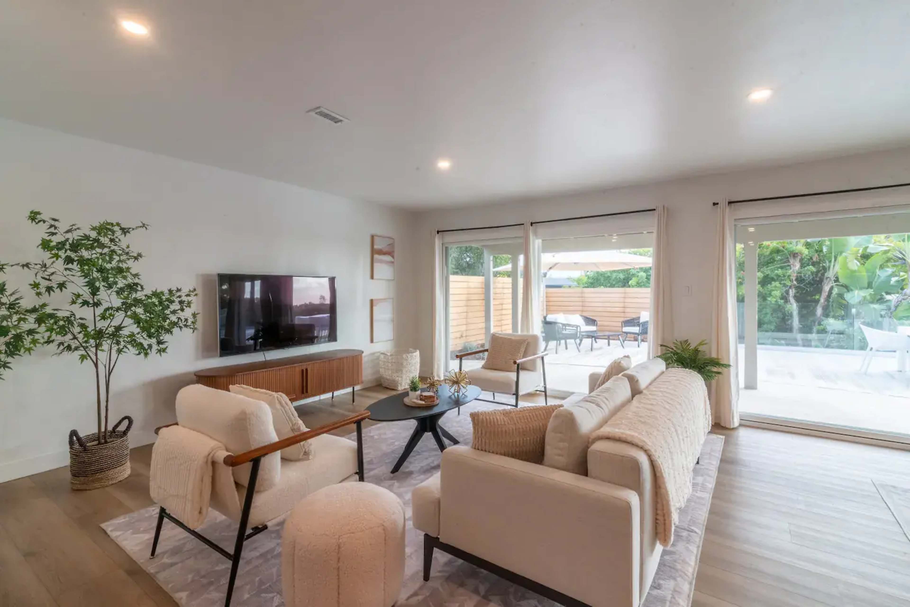 A modern living room features a white sofa set, a wooden TV console, and large sliding glass doors that open to an outdoor patio area.