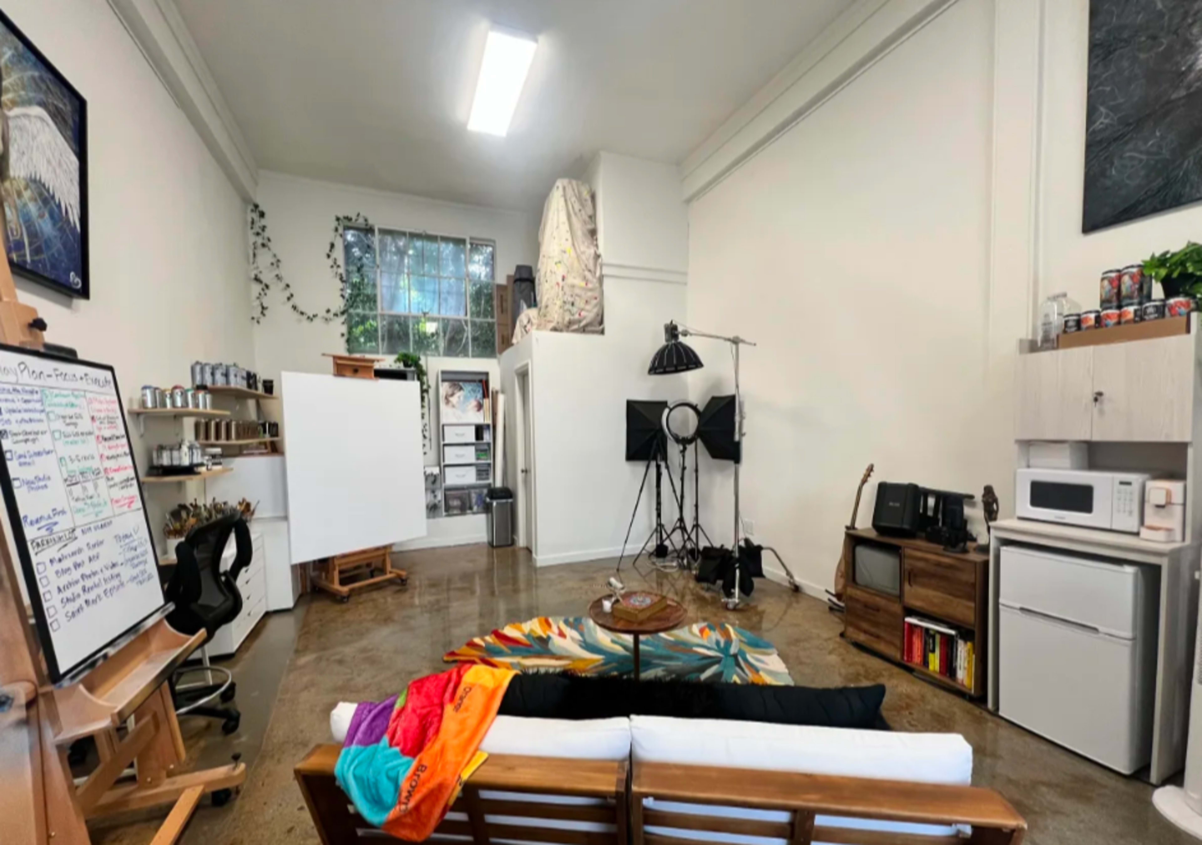 The image depicts a spacious, well-lit room featuring a sofa with colorful cushions, a large rug, an easel, two stands with equipment, and shelves stocked with various items.