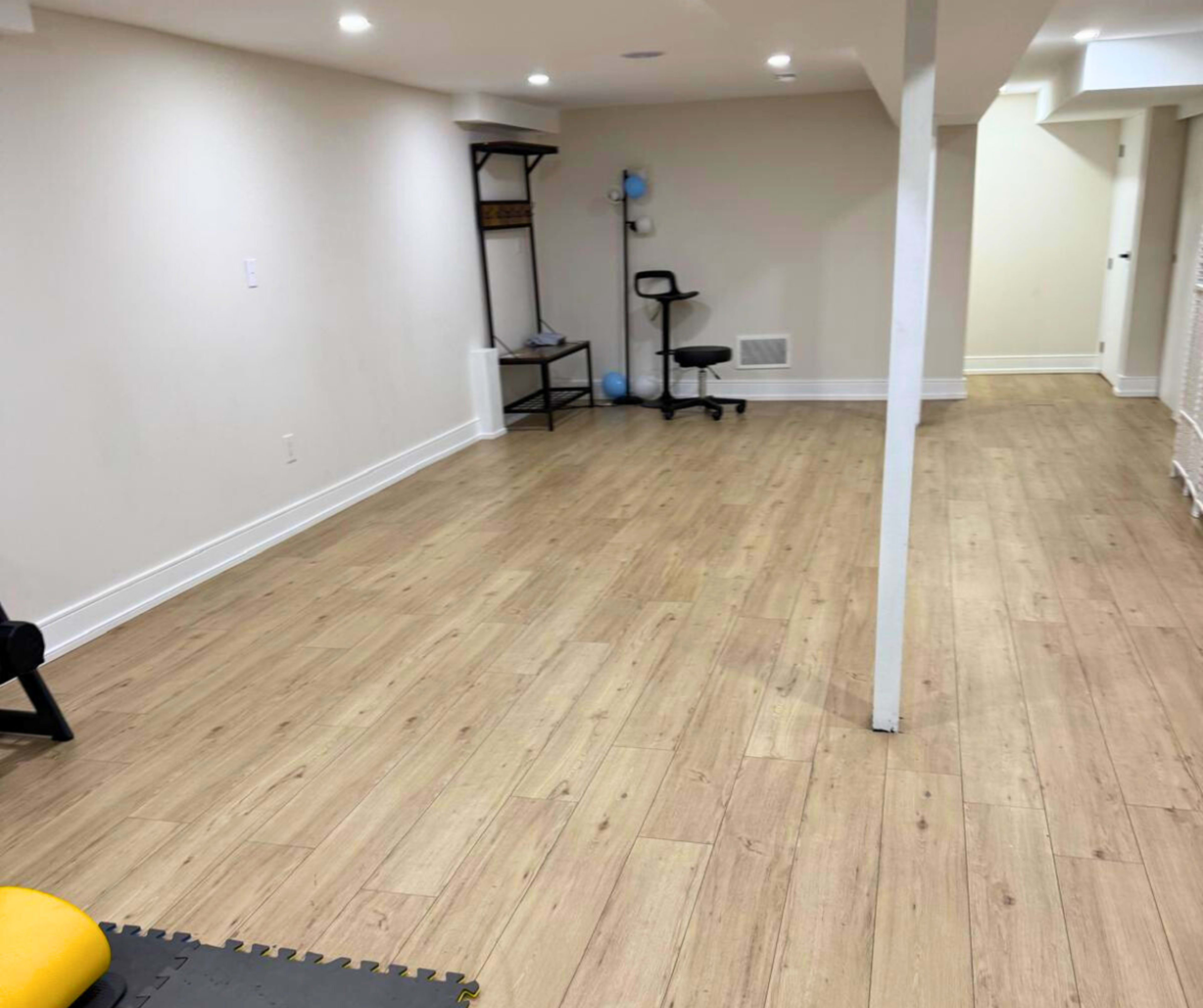 The image shows a spacious, well-lit basement with wooden flooring, featuring minimal furniture and a small chair in the corner.