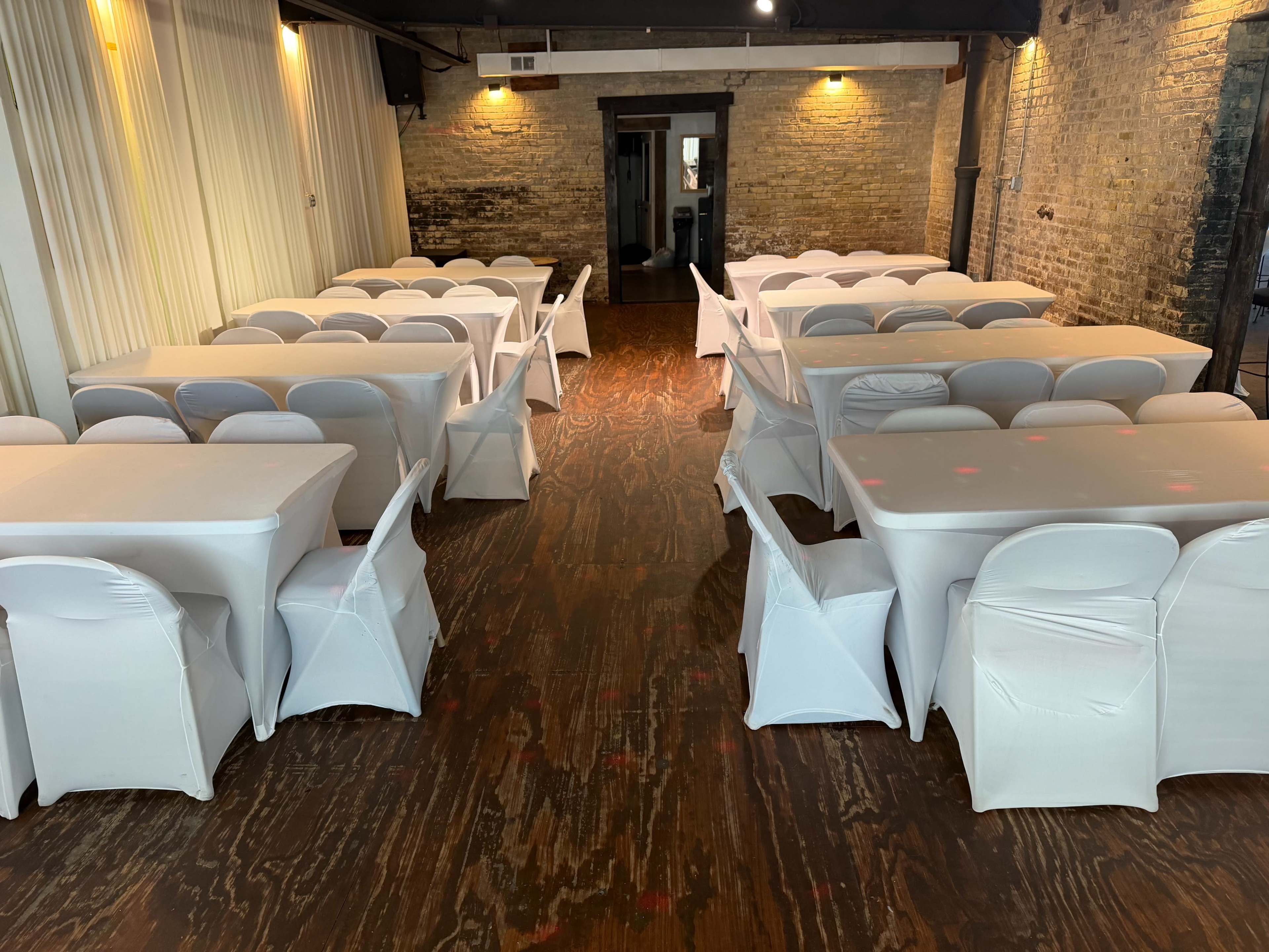 The image shows a spacious event venue with white tablecloths covering several tables arranged in a grid pattern, surrounded by white chairs on a wooden floor.