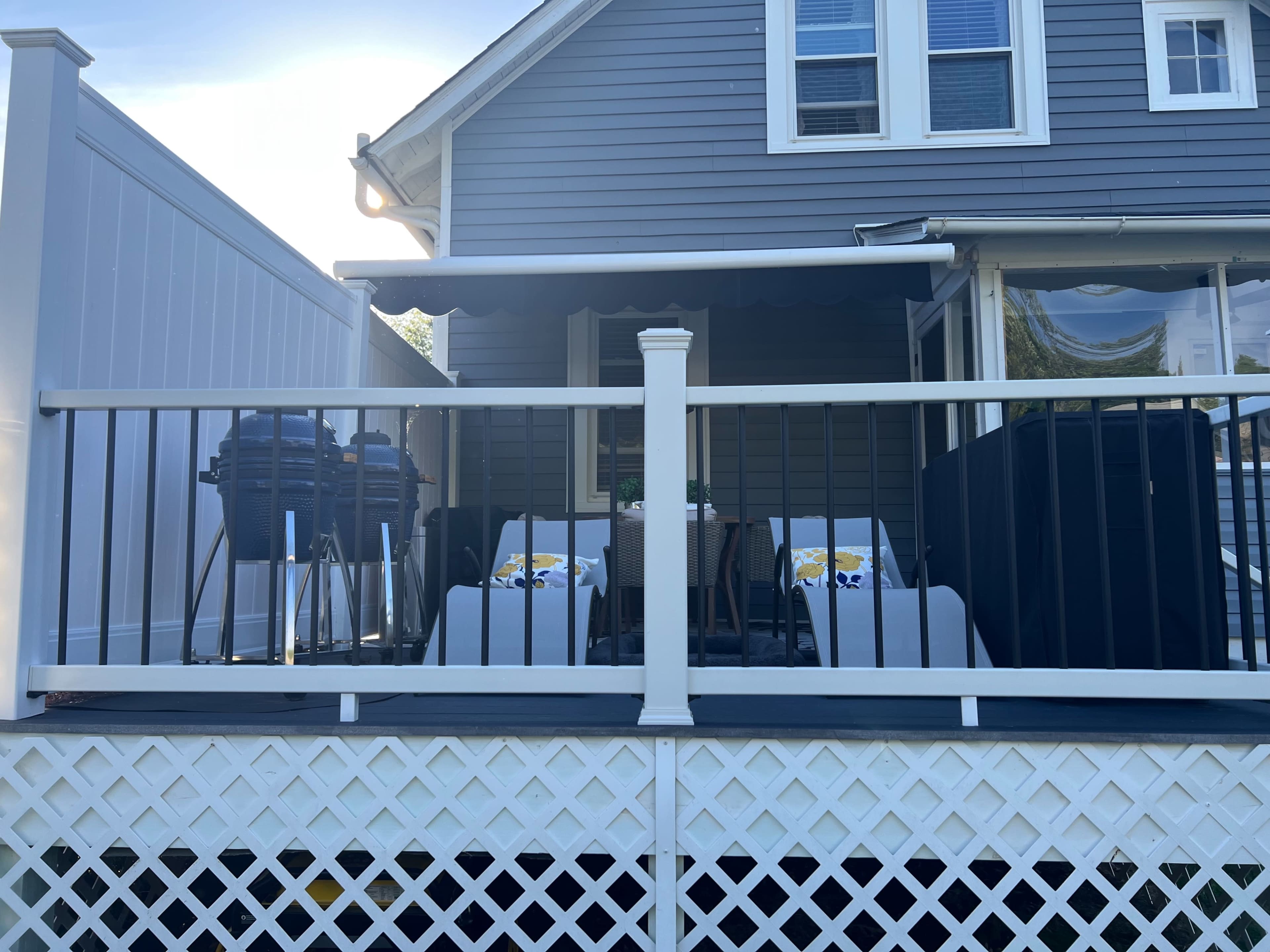 The image shows a backyard deck with two lounge chairs, a table, and a grill, all situated against the side of a gray house.