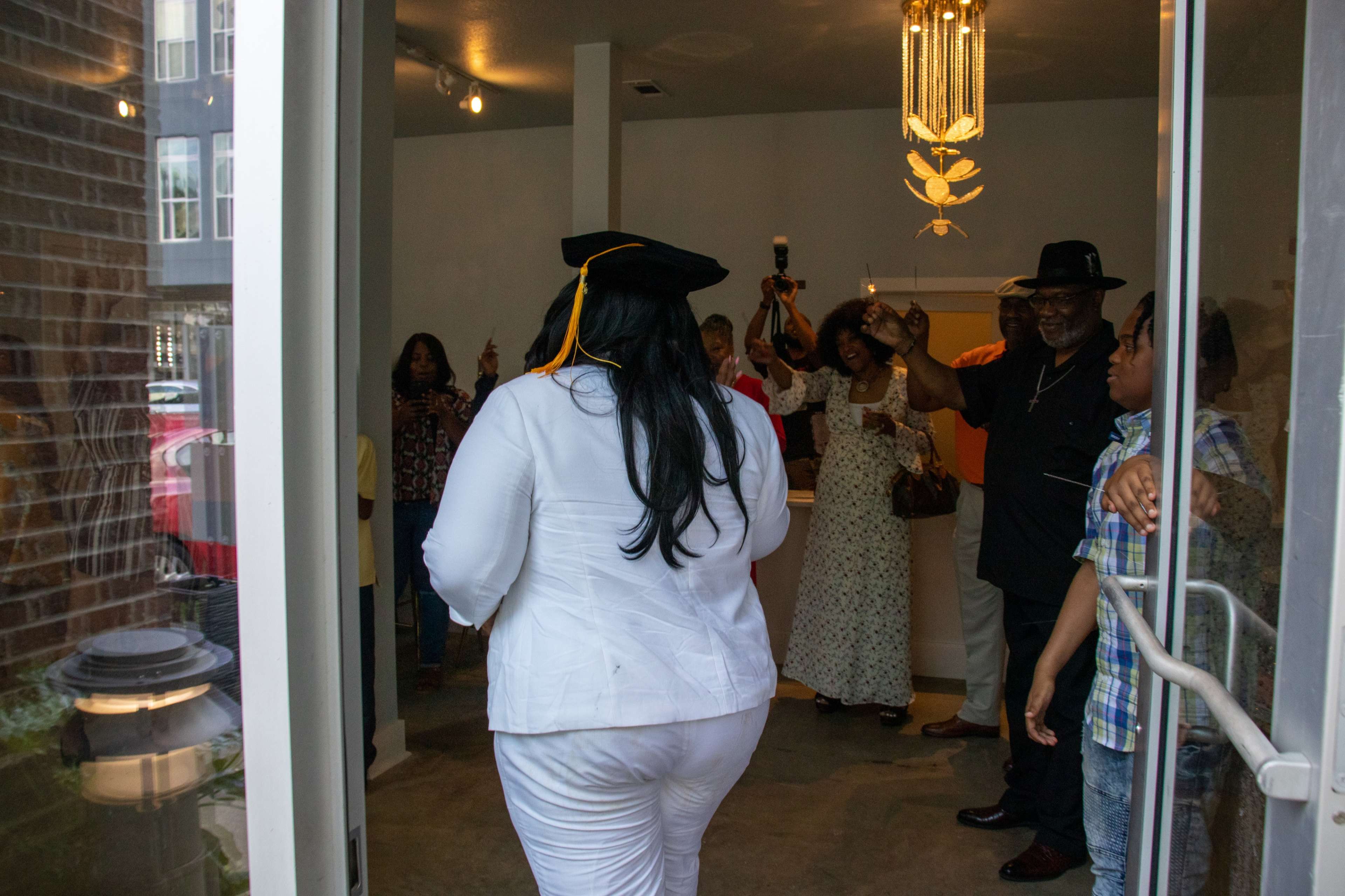 A woman in a white outfit and graduation cap walks through a doorway while a group of people celebrate inside.