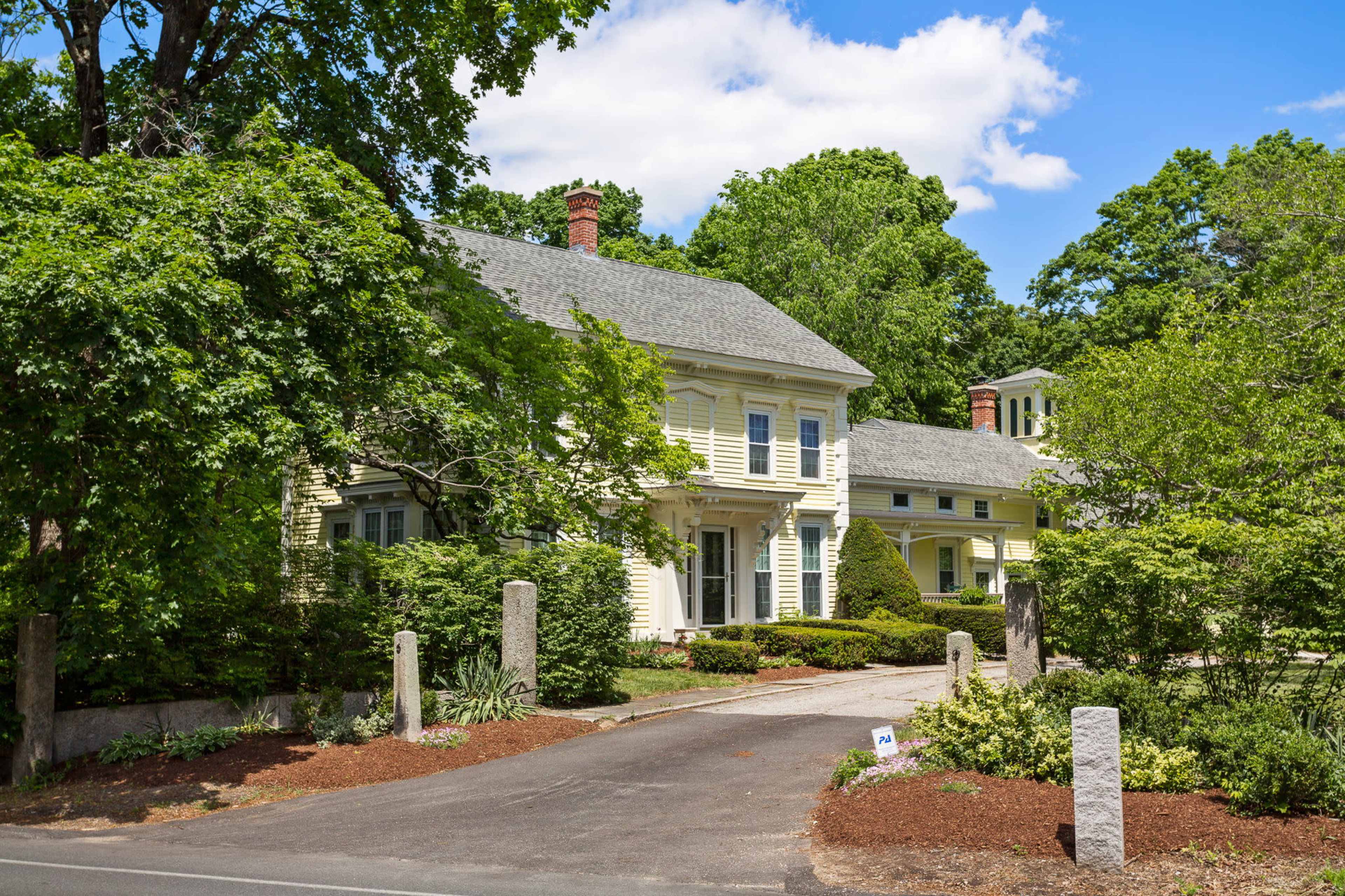 A large, yellow, two-story house surrounded by trees and greenery, with a paved driveway leading to the entrance.