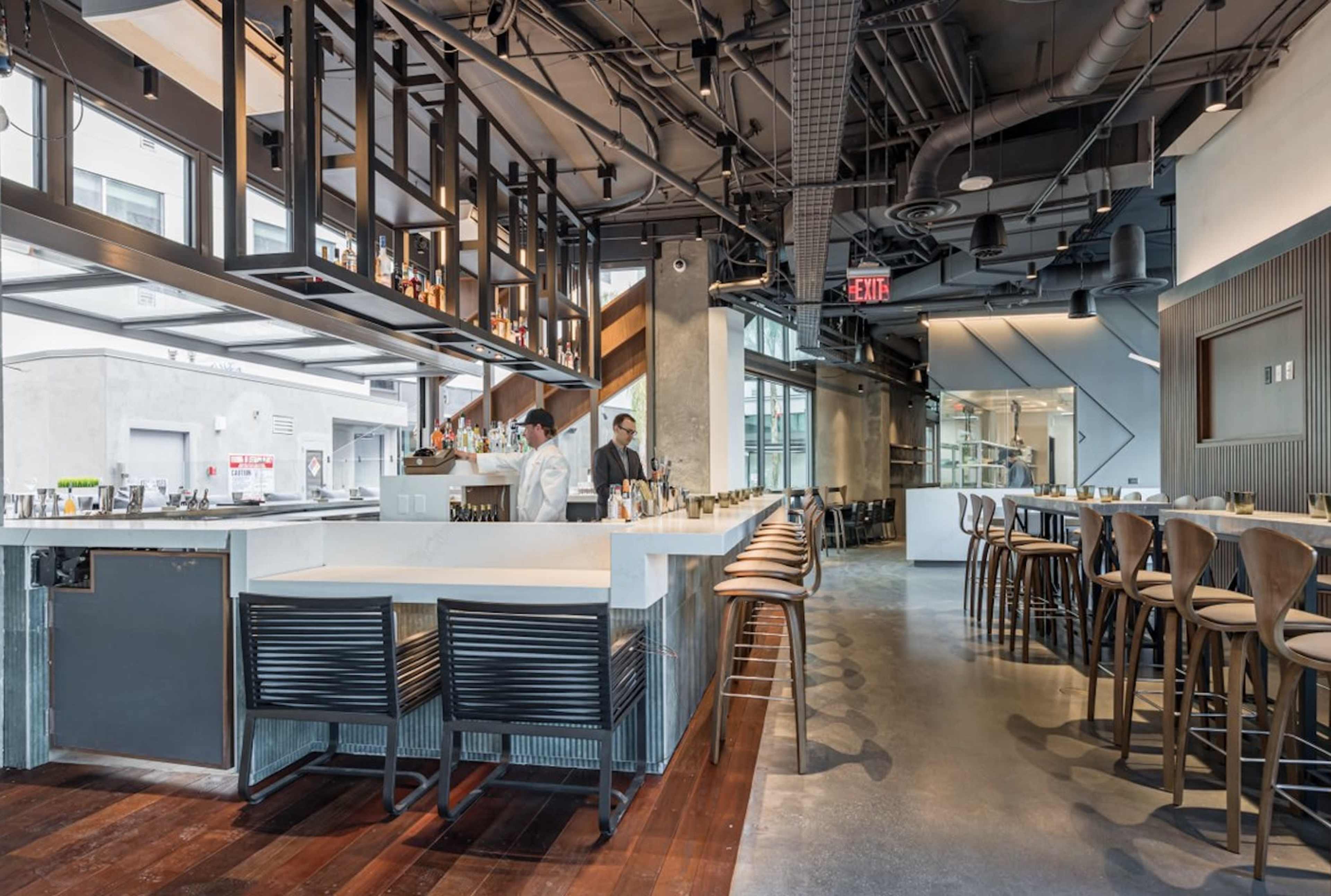 The image shows a modern restaurant interior with a bar area, high stools, and two chefs working behind the counter.