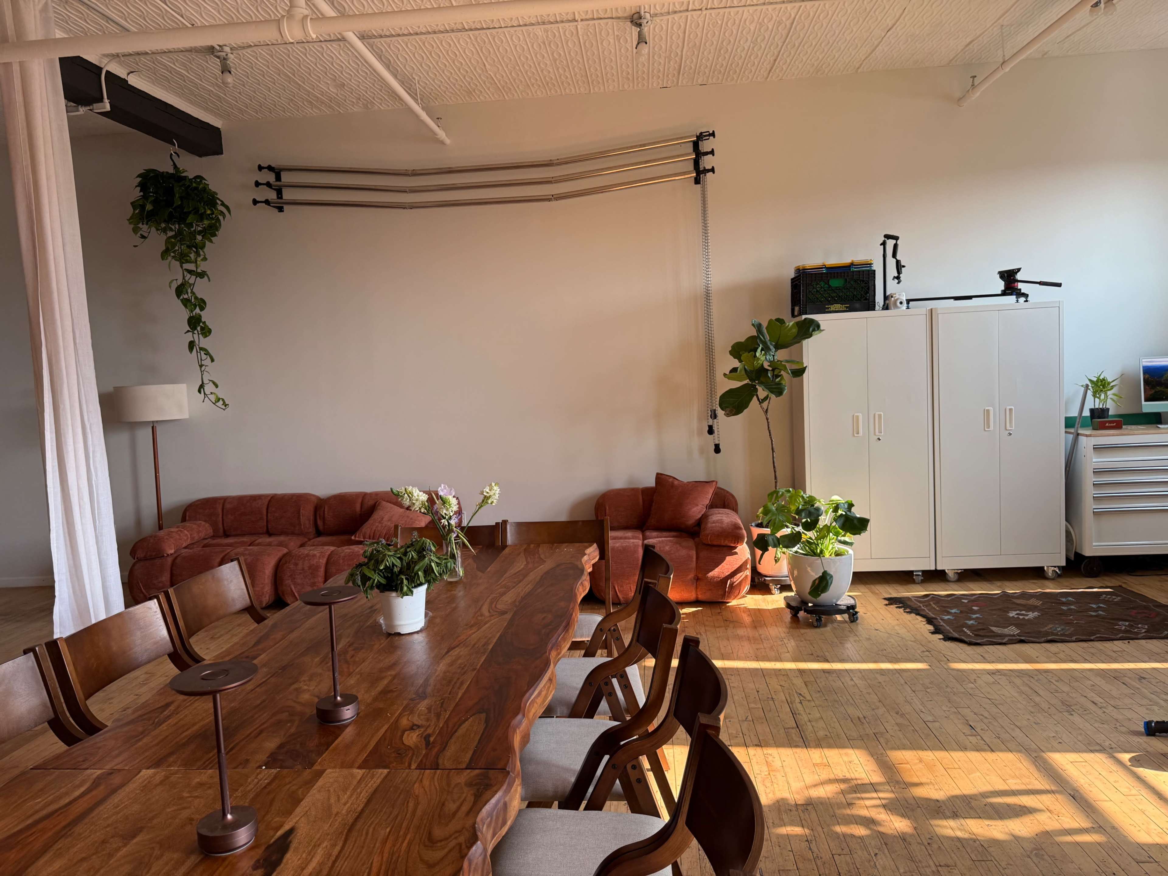 The image shows a bright, minimalist interior space featuring a wooden dining table, a red sofa, houseplants, and storage cabinets along the wall.
