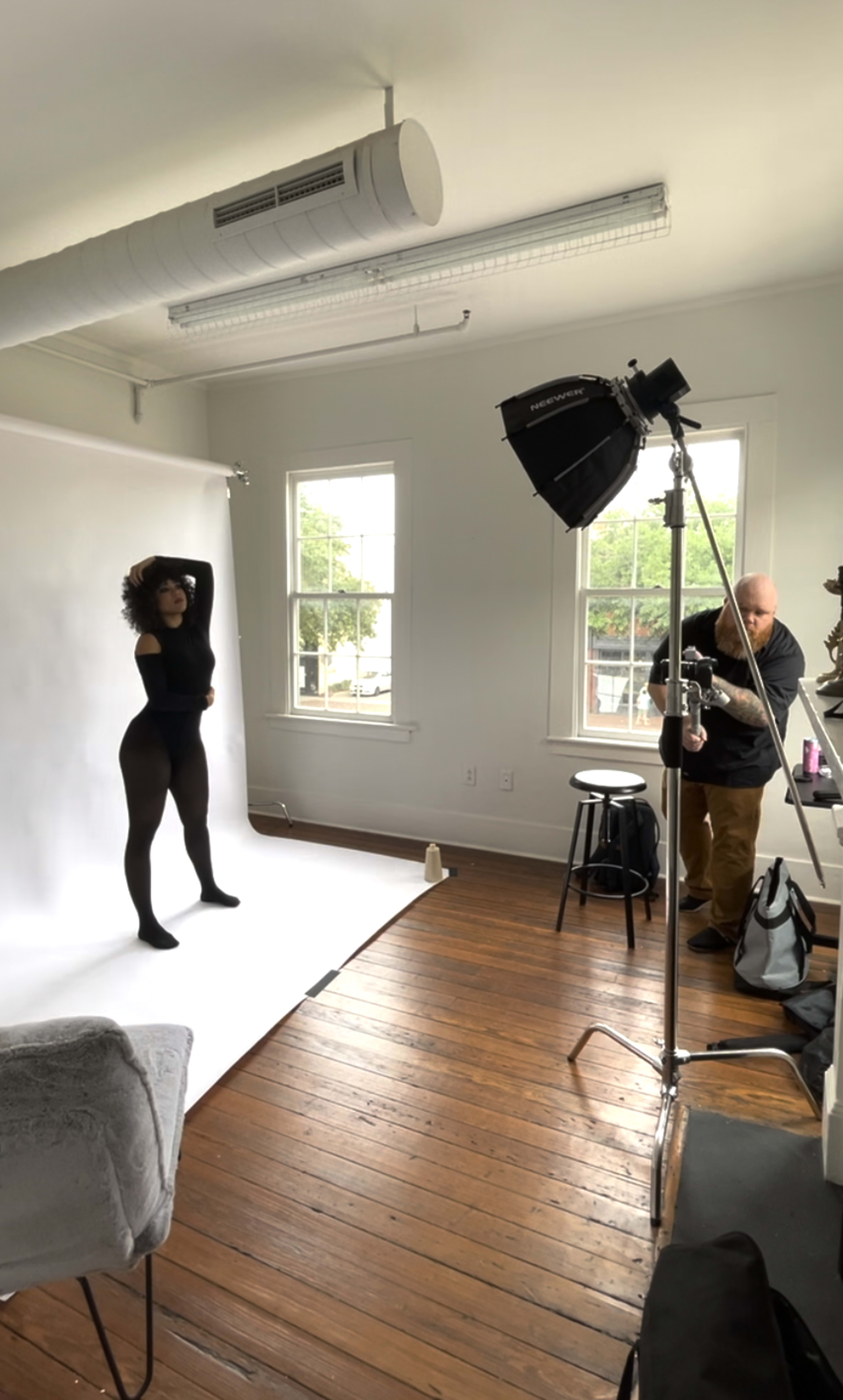 A model poses in a black outfit against a white backdrop while a photographer adjusts lighting in a well-lit studio.