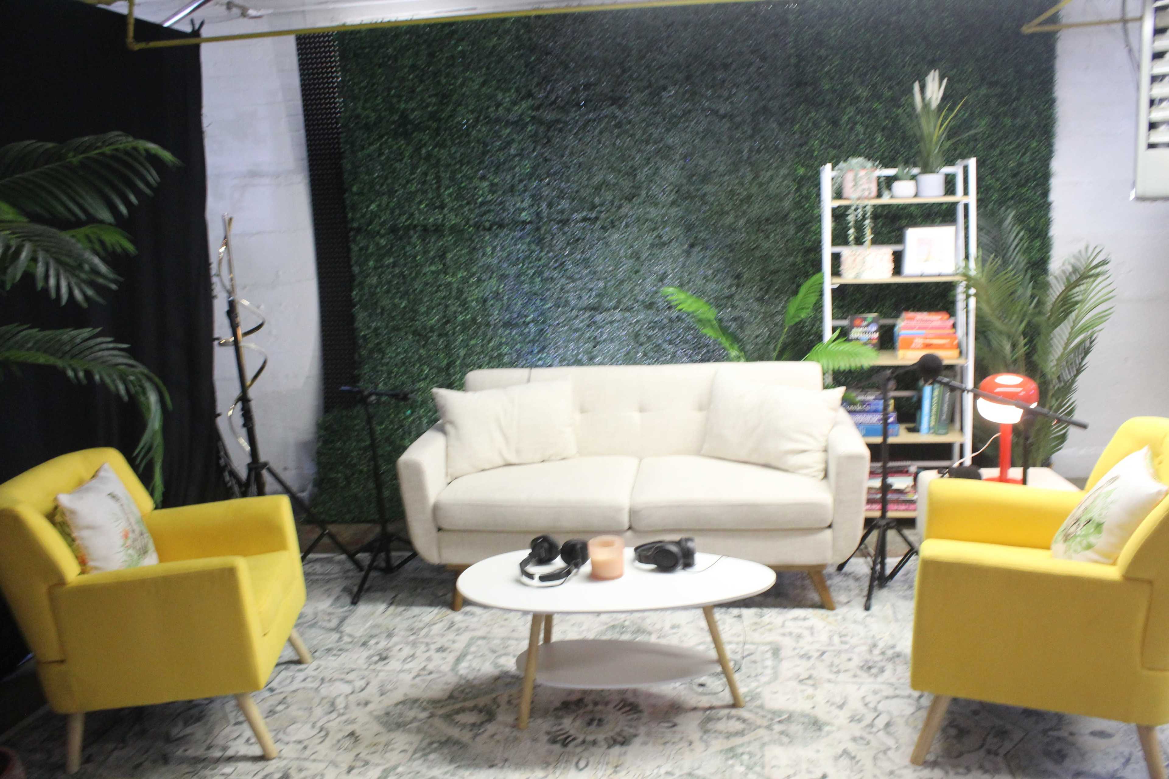The image shows a modern indoor space featuring a light-colored sofa and two yellow armchairs arranged around a white coffee table, with a backdrop of green foliage and a shelf filled with books and decorative items.