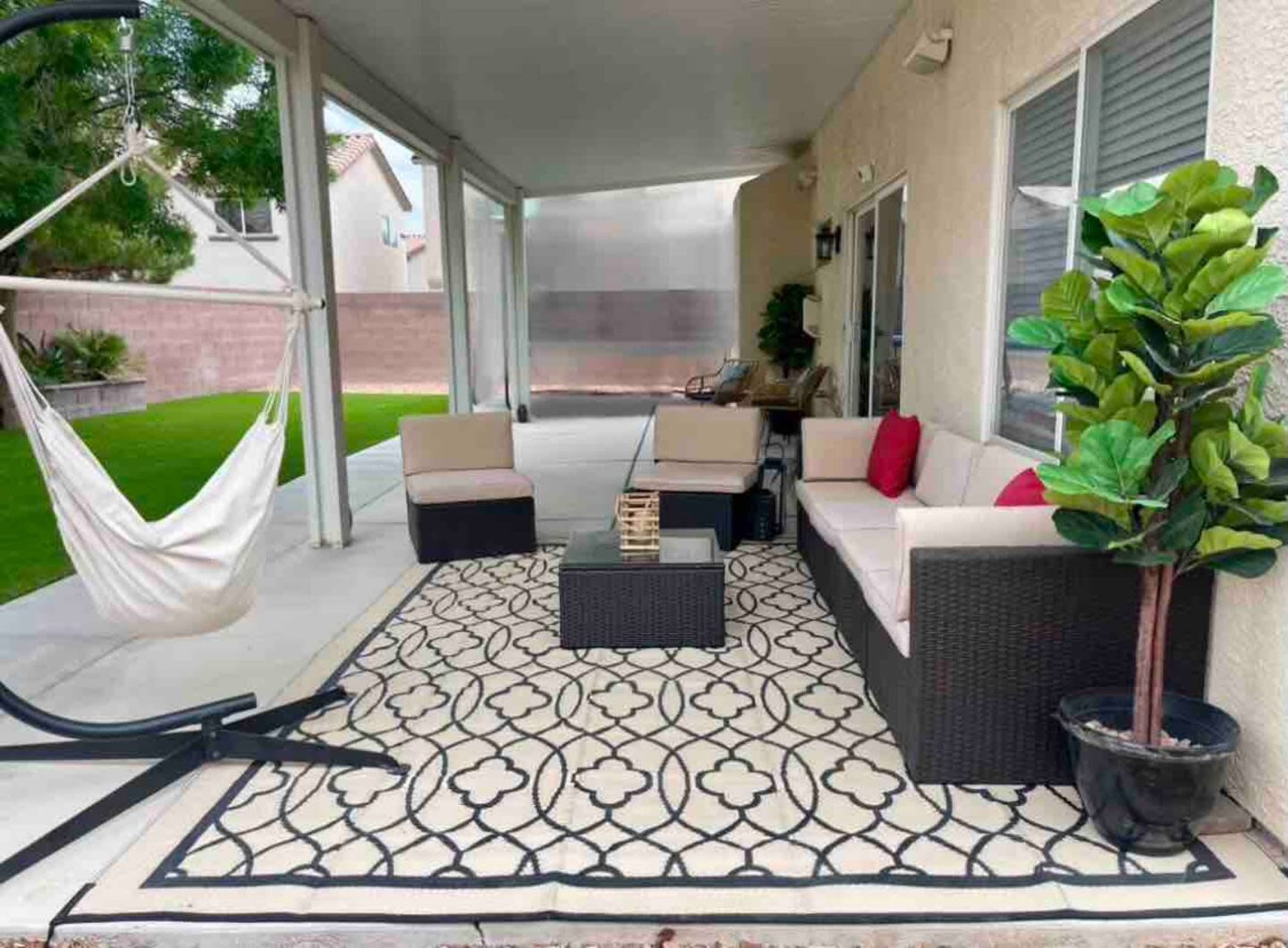 The image shows a patio area with a seating arrangement consisting of a sofa and chairs, a small coffee table, a hammock, and decorative plants on a patterned rug.