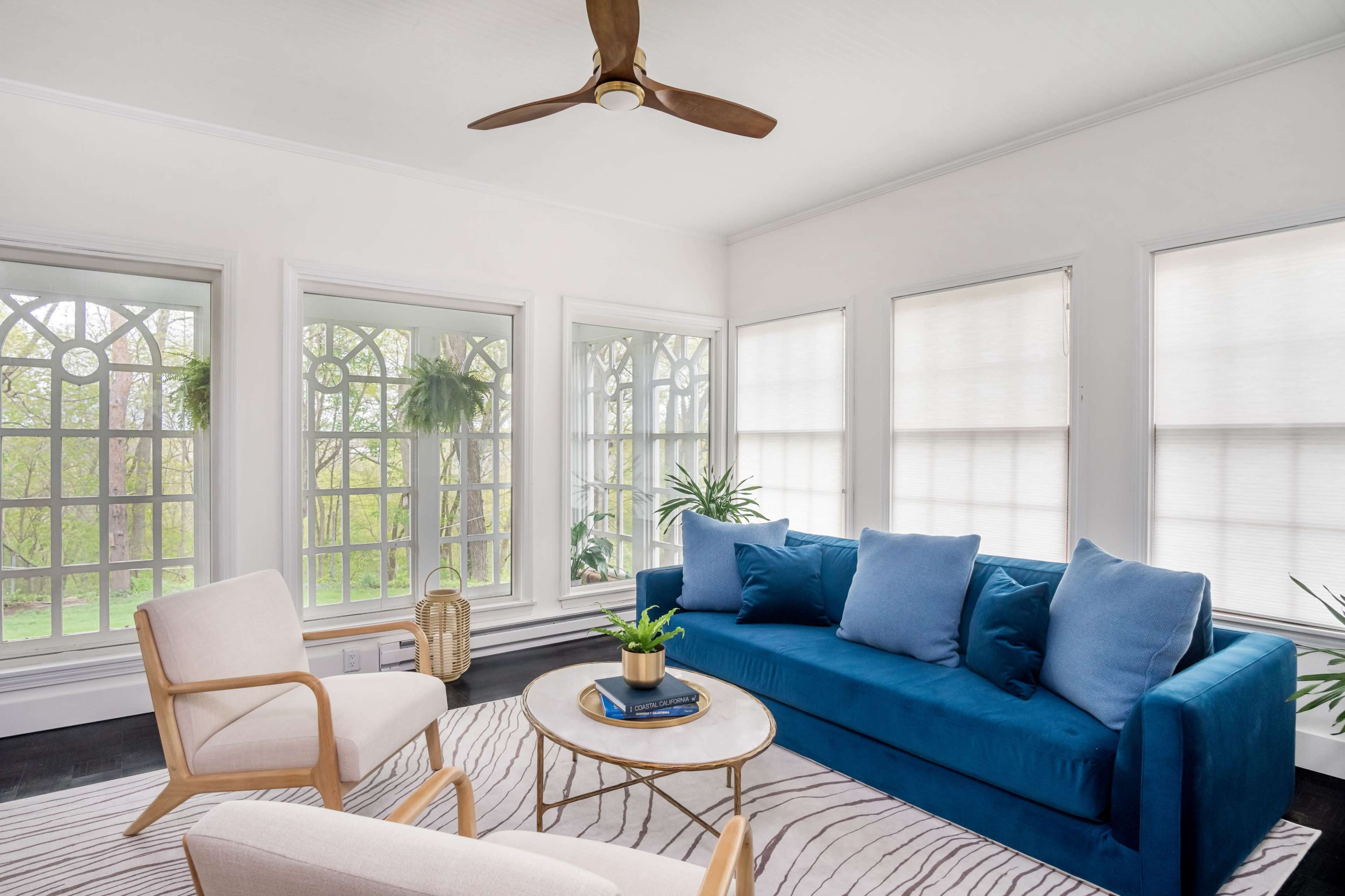 A bright sunroom features a blue sofa, two cream chairs, a round coffee table, and large windows surrounded by greenery.