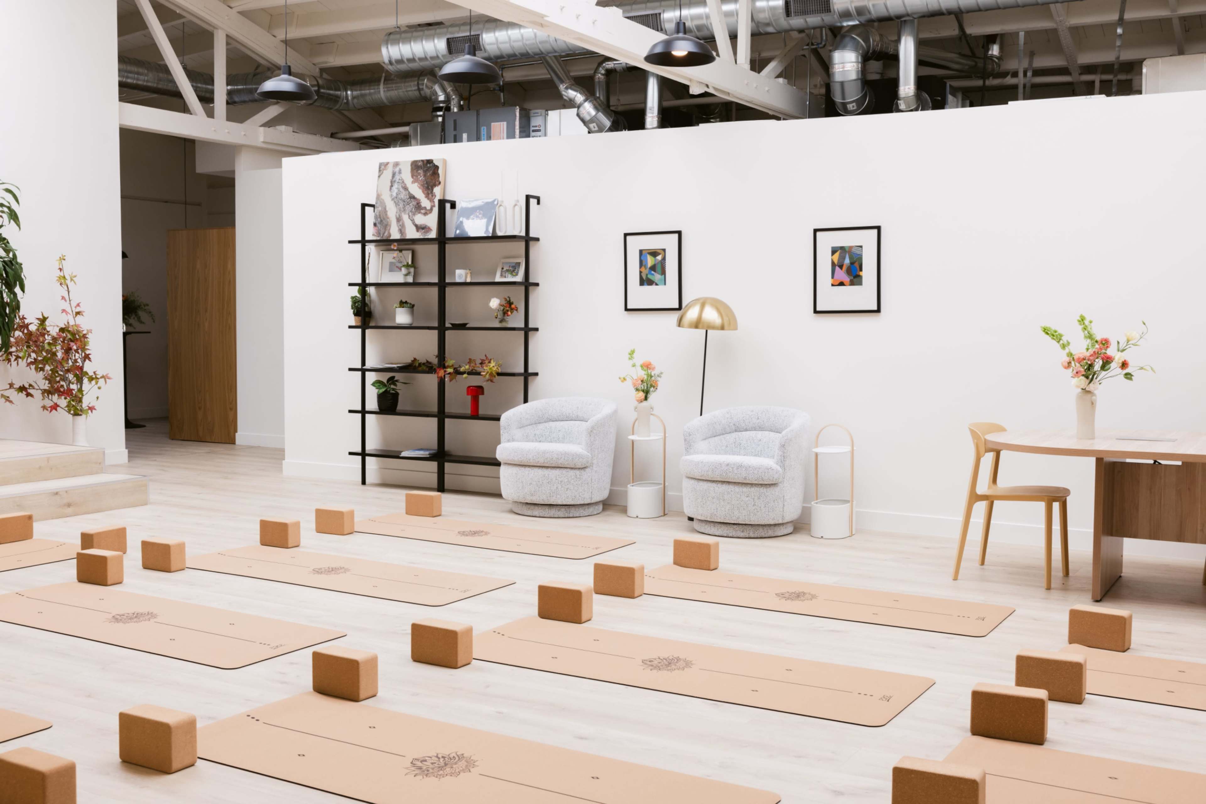 The image depicts a yoga studio with neatly arranged mat spaces, wooden blocks, a cozy seating area, and decorative shelves along a white wall.