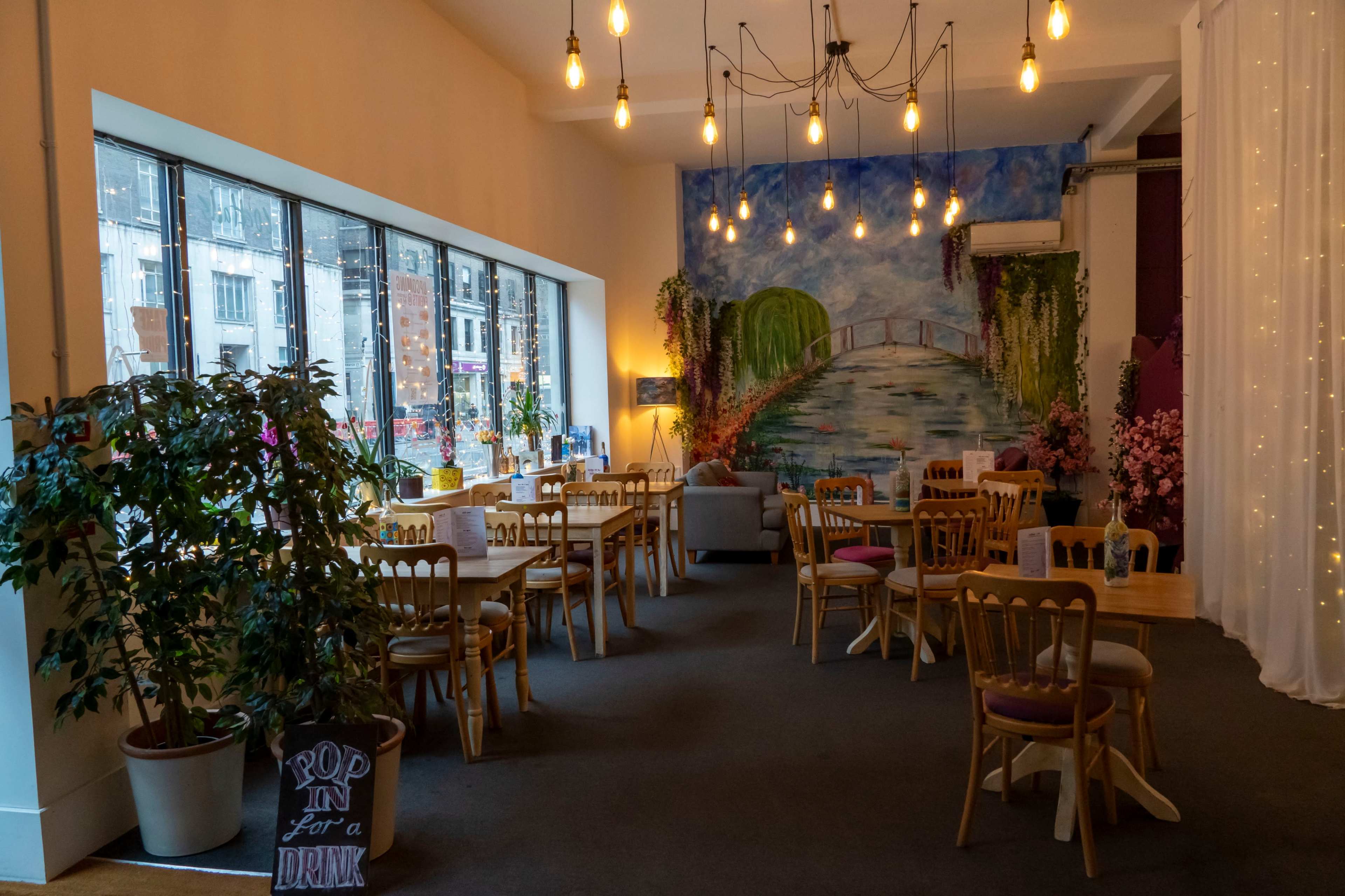 The image shows a cozy cafe interior with wooden tables and chairs, large windows, and a mural of a river landscape on the wall.
