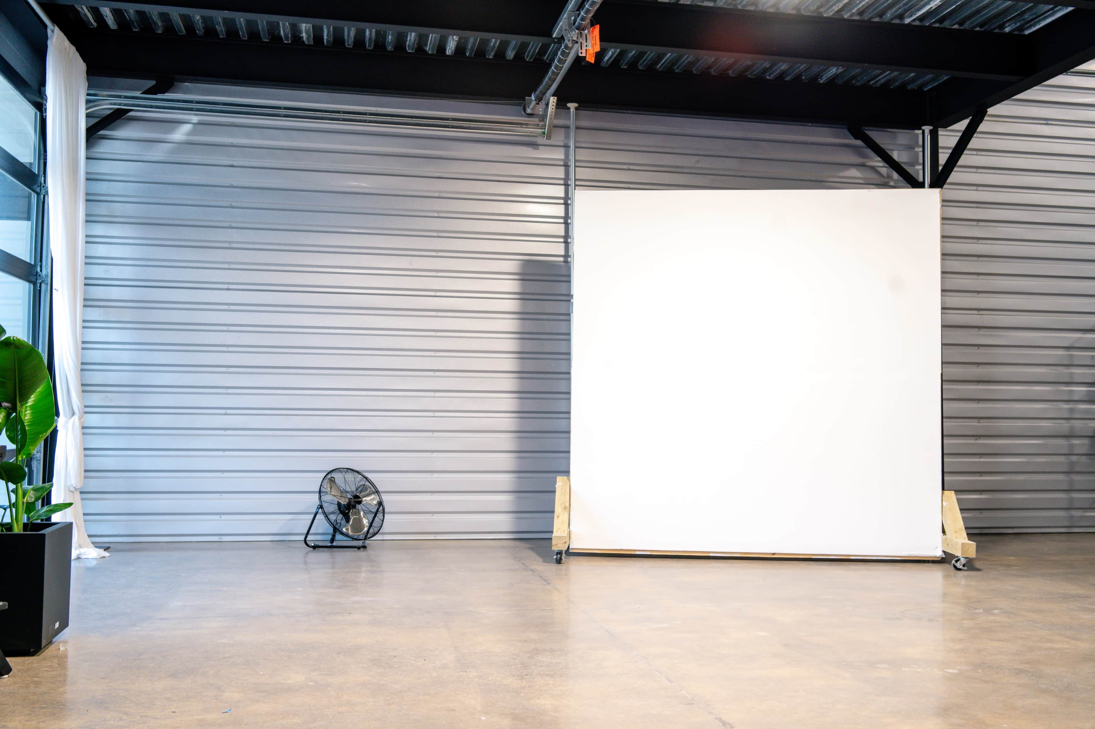A large blank white canvas stands on wooden supports in an industrial space with a concrete floor and a small fan in the corner.