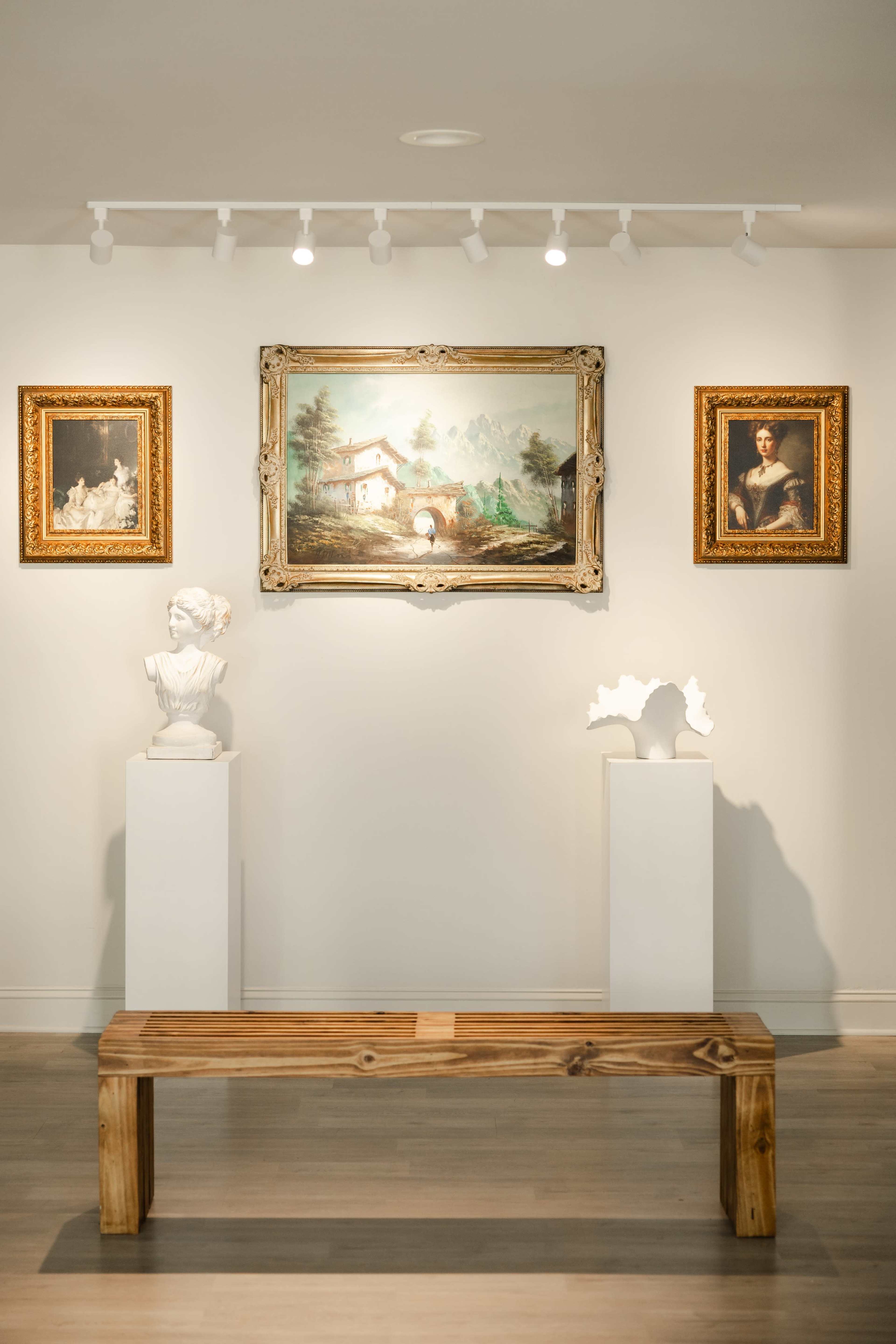 The image shows an art gallery featuring three framed paintings on the wall and two white pedestals displaying sculptures, with a wooden bench positioned in front.