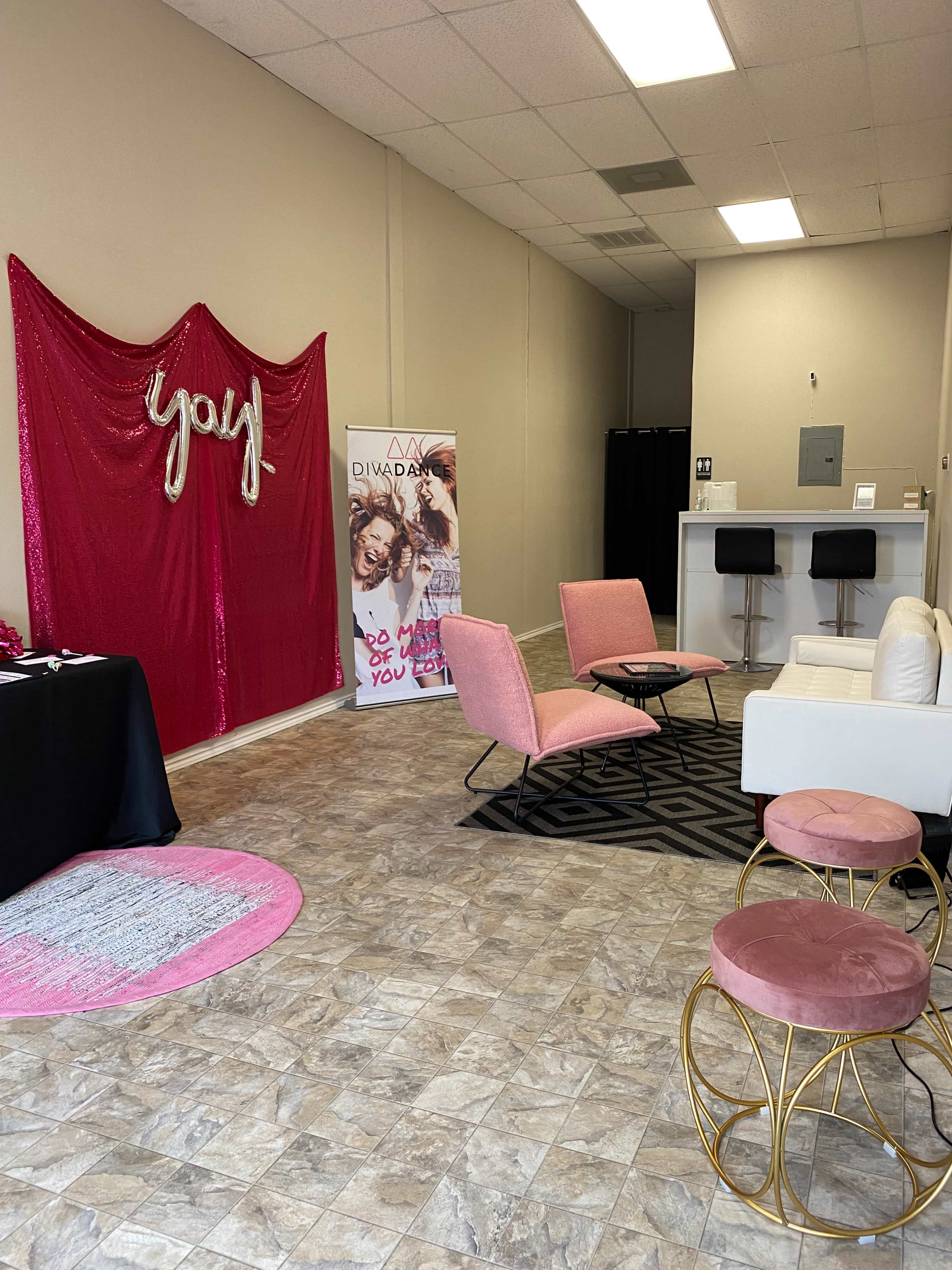 A spacious, well-lit interior featuring pink lounge chairs, a white sofa, and a large decorative wall with the word "you," alongside promotional banners and a black table.