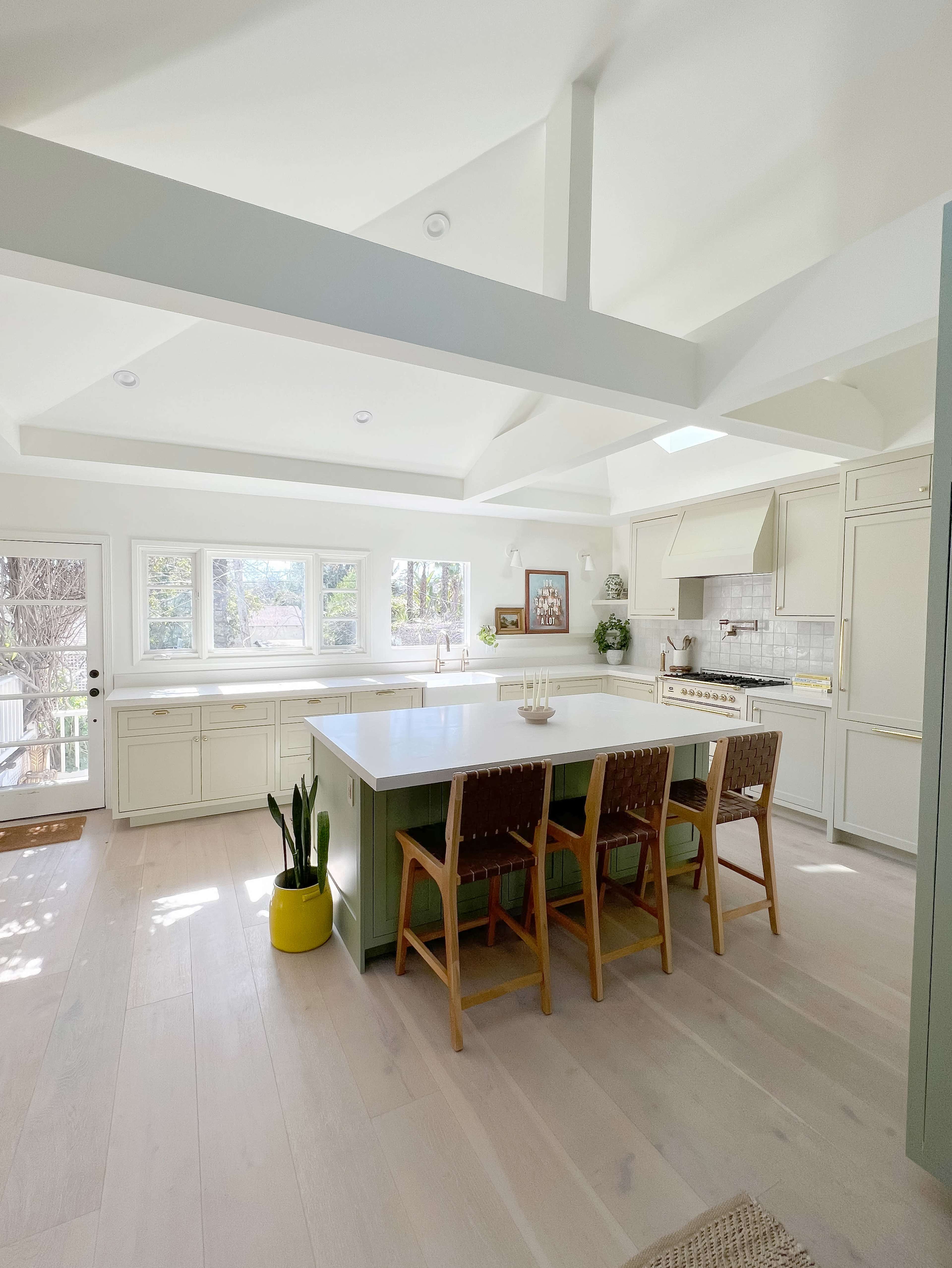 A modern kitchen features a large island with wooden stools, bright lighting, and windows overlooking a natural landscape.