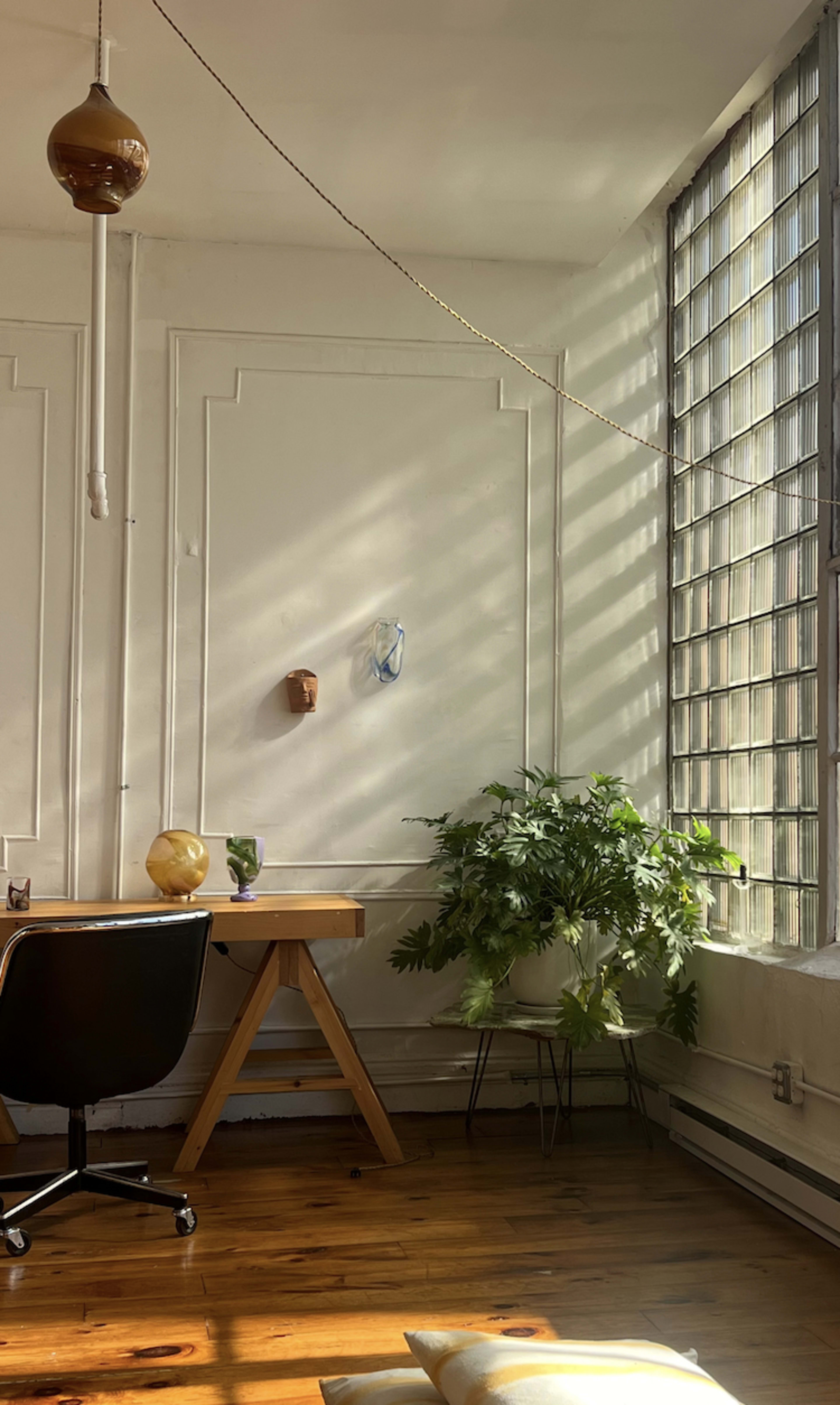A well-lit room features a desk with a black chair, a plant beside a glass block window, and decorative wall accents.