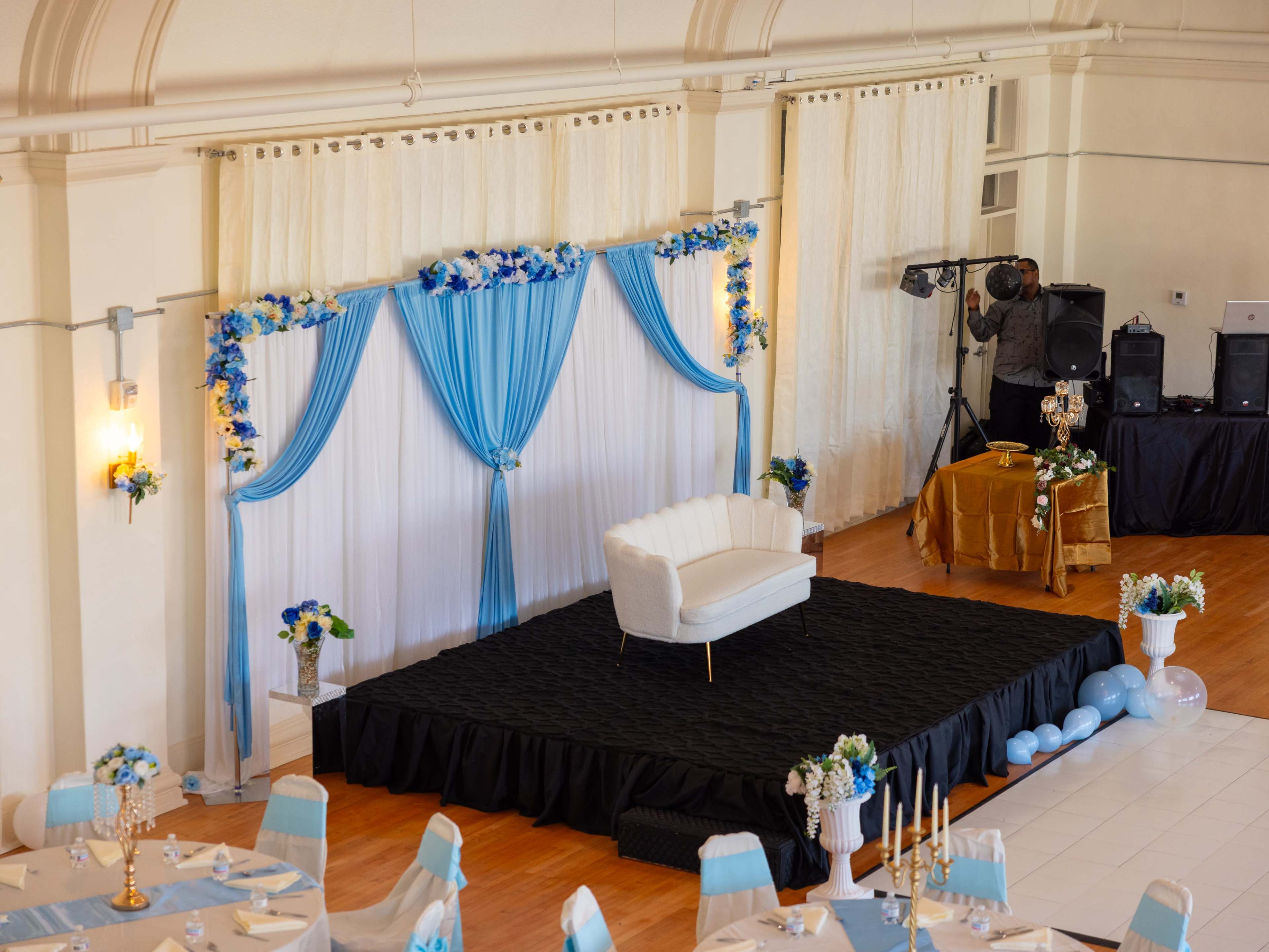 The image shows a decorated event space with a white couch on a raised black platform, surrounded by blue and white drapery, floral arrangements, and tables set for a gathering.