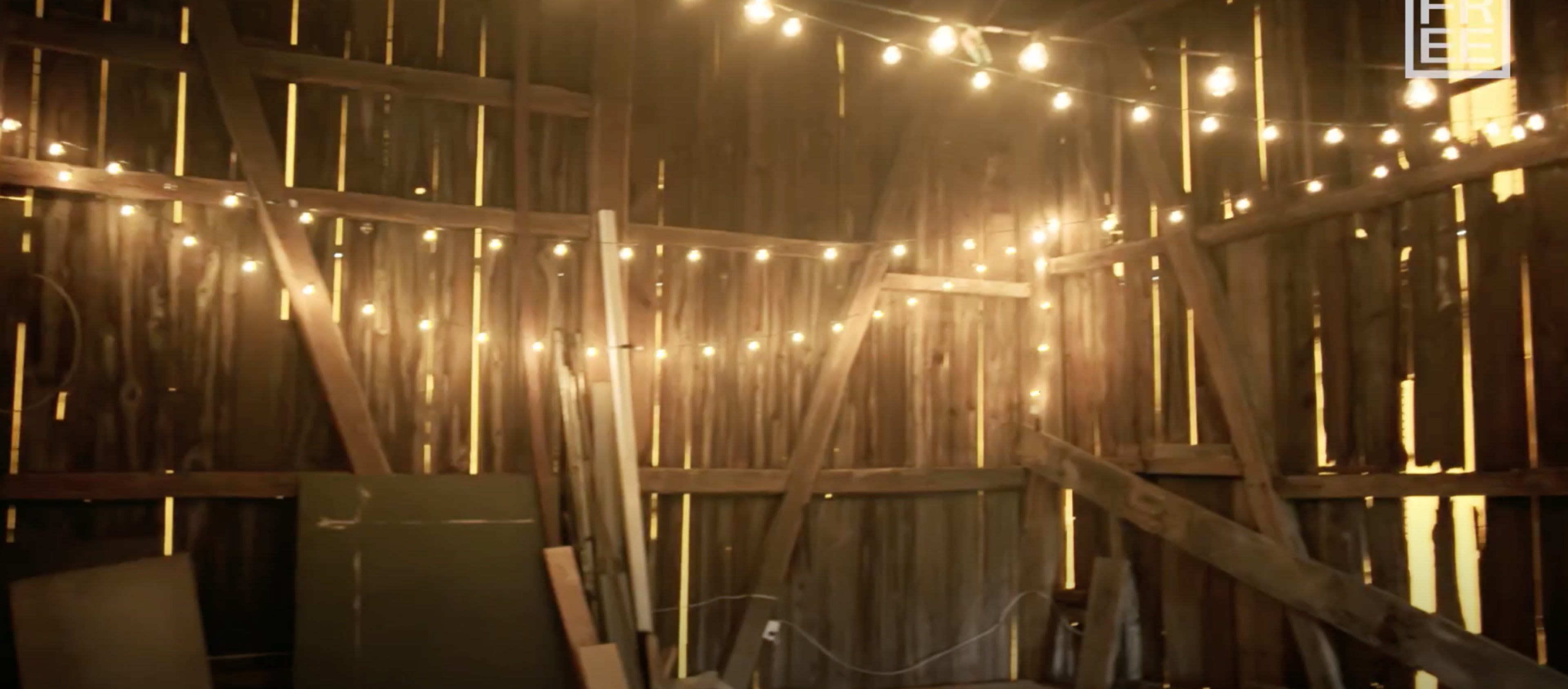 The interior of a wooden barn features string lights illuminating the space and various pieces of wood and furniture stored along the walls.