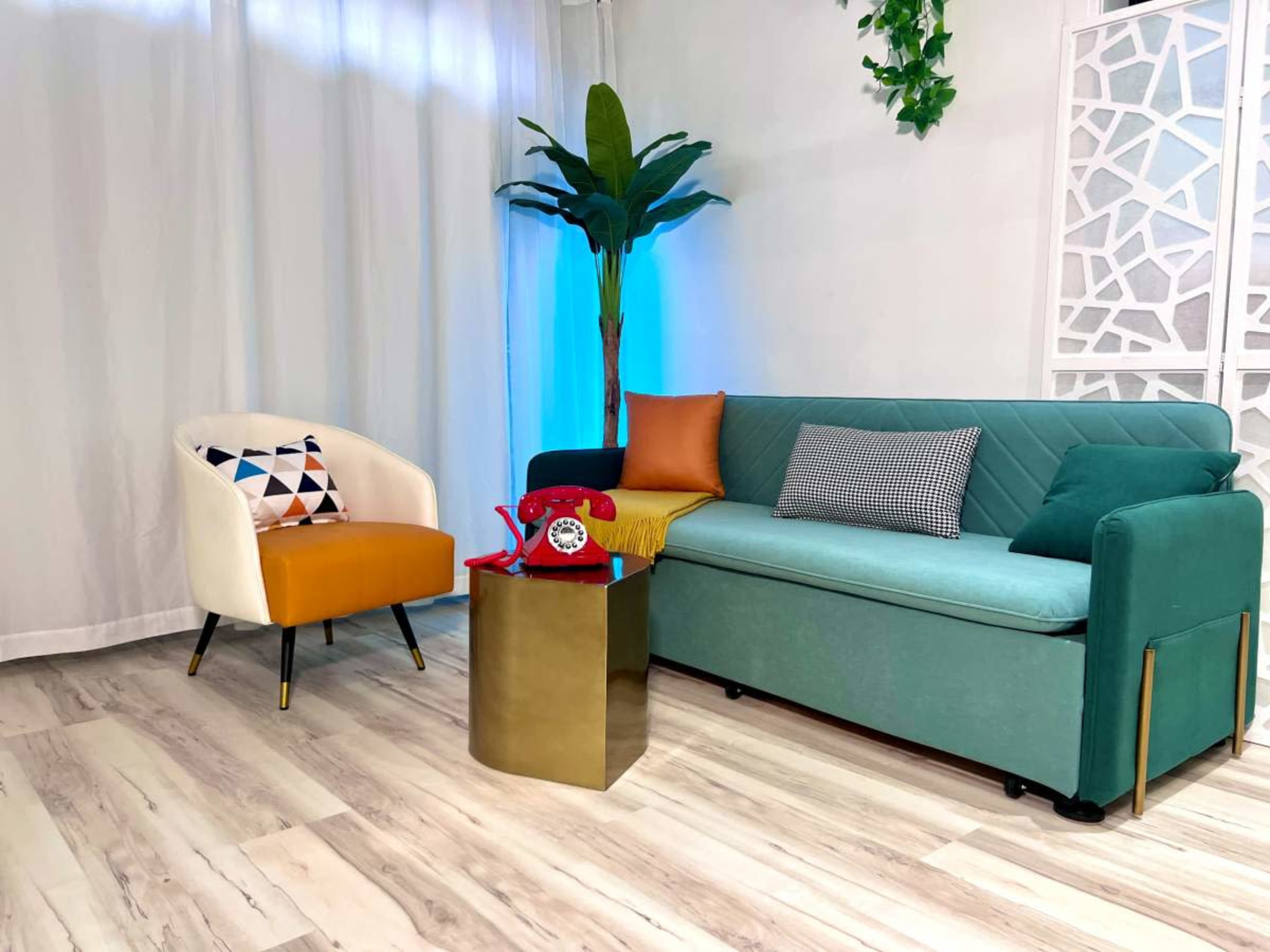 The image shows a modern seating area featuring a turquoise couch, a cream-colored accent chair with orange upholstery, and a gold side table with a red rotary phone on it, against a backdrop of white curtains and greenery.