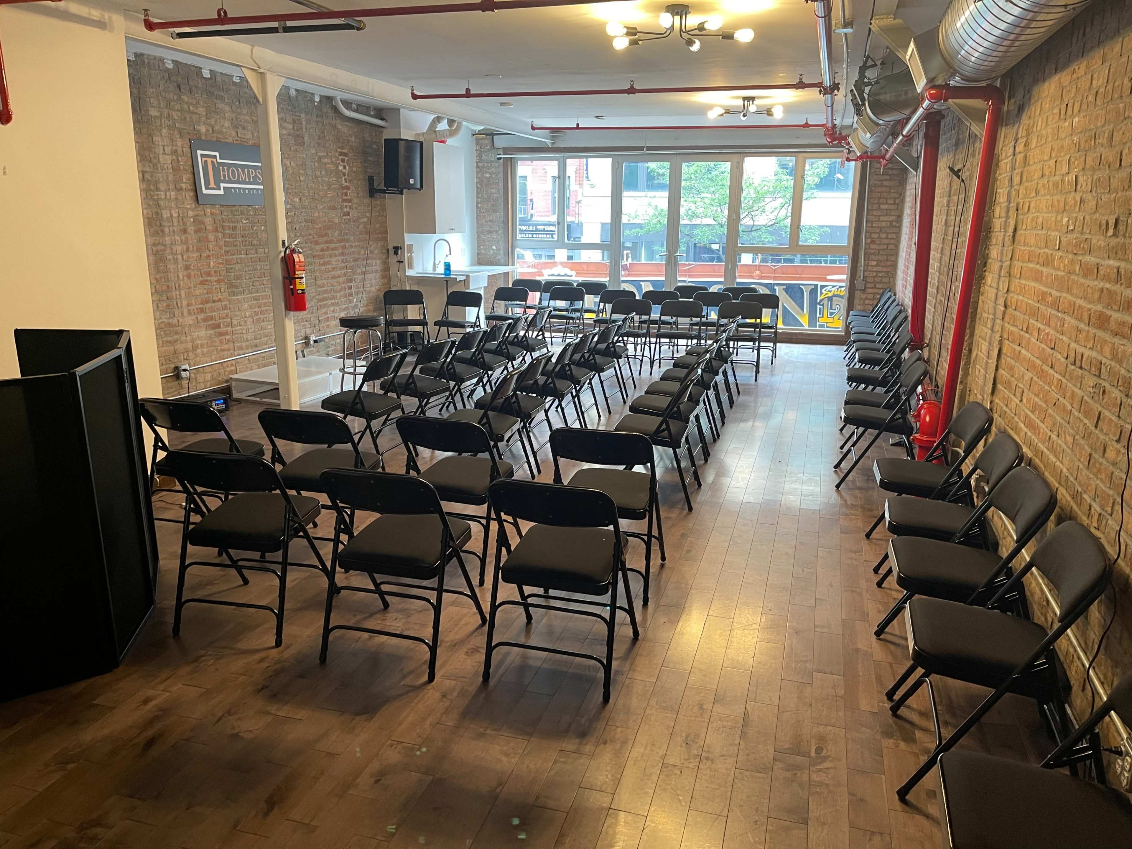 The room features multiple rows of black folding chairs arranged facing a large window, with exposed brick walls and wooden flooring.