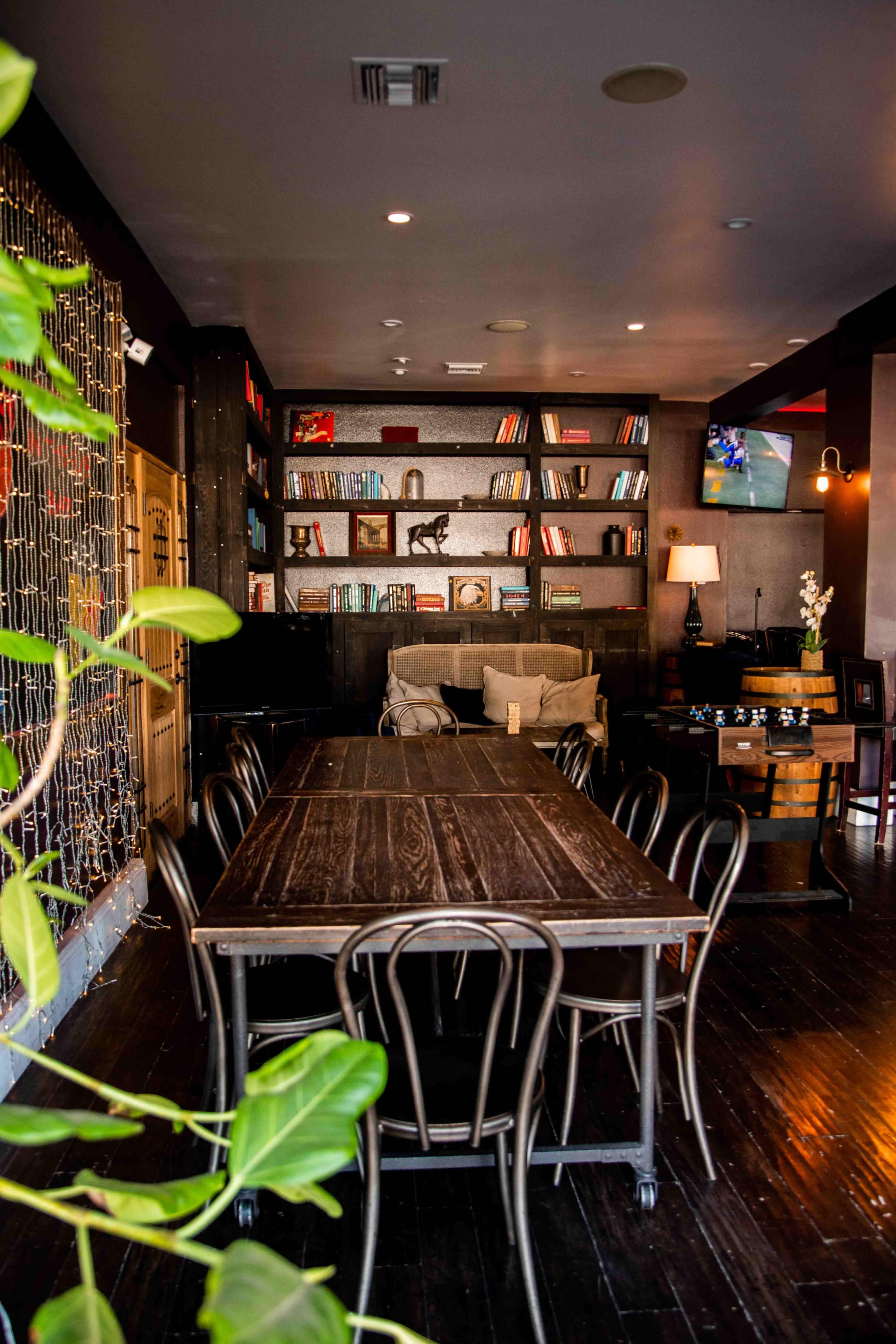 The image shows a cozy café interior featuring a dark wooden table surrounded by chairs, with shelves filled with books and a television in the background.
