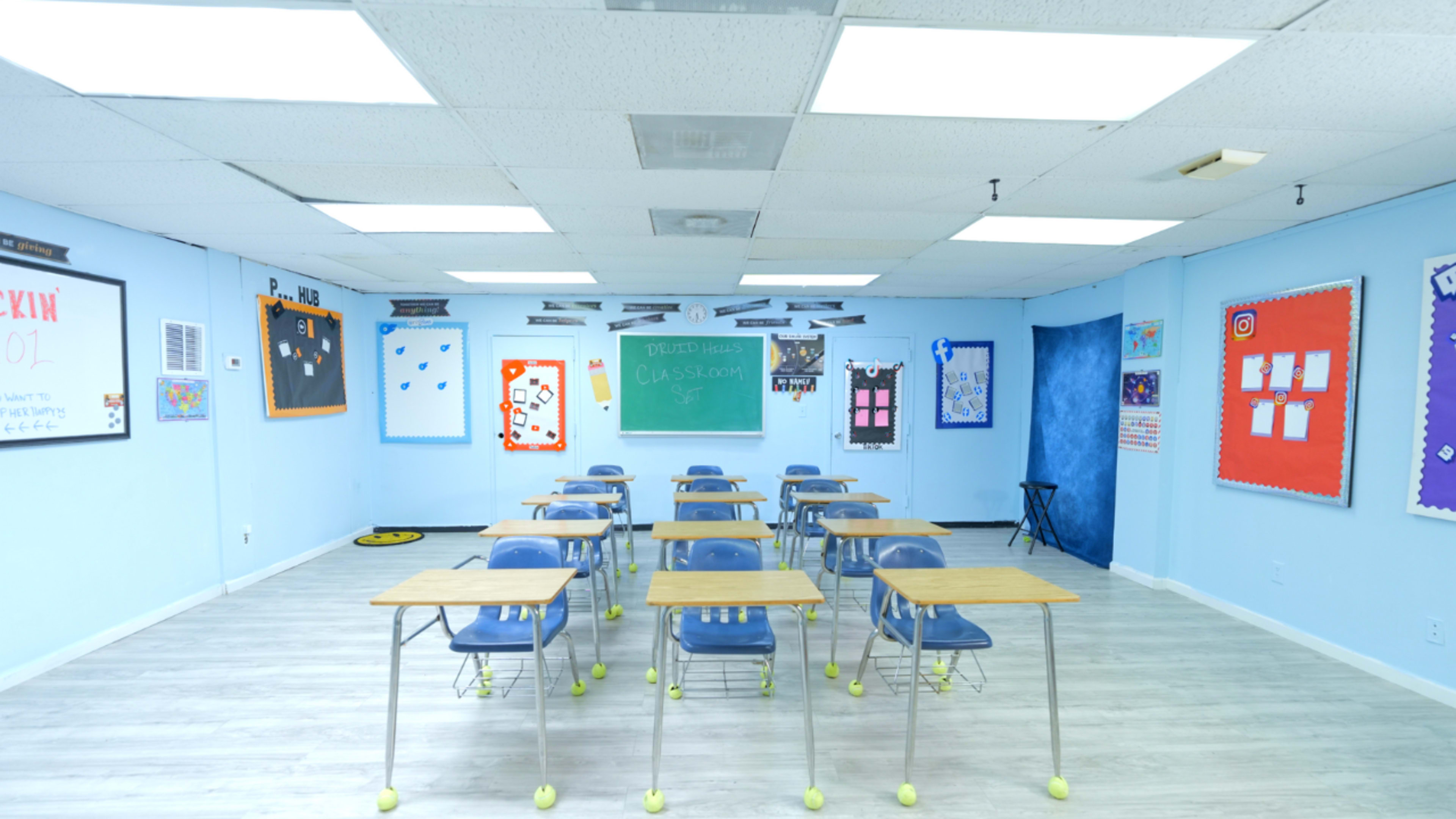 The image shows a classroom with several desks arranged in rows, a chalkboard at the front, and colorful educational posters on the walls.