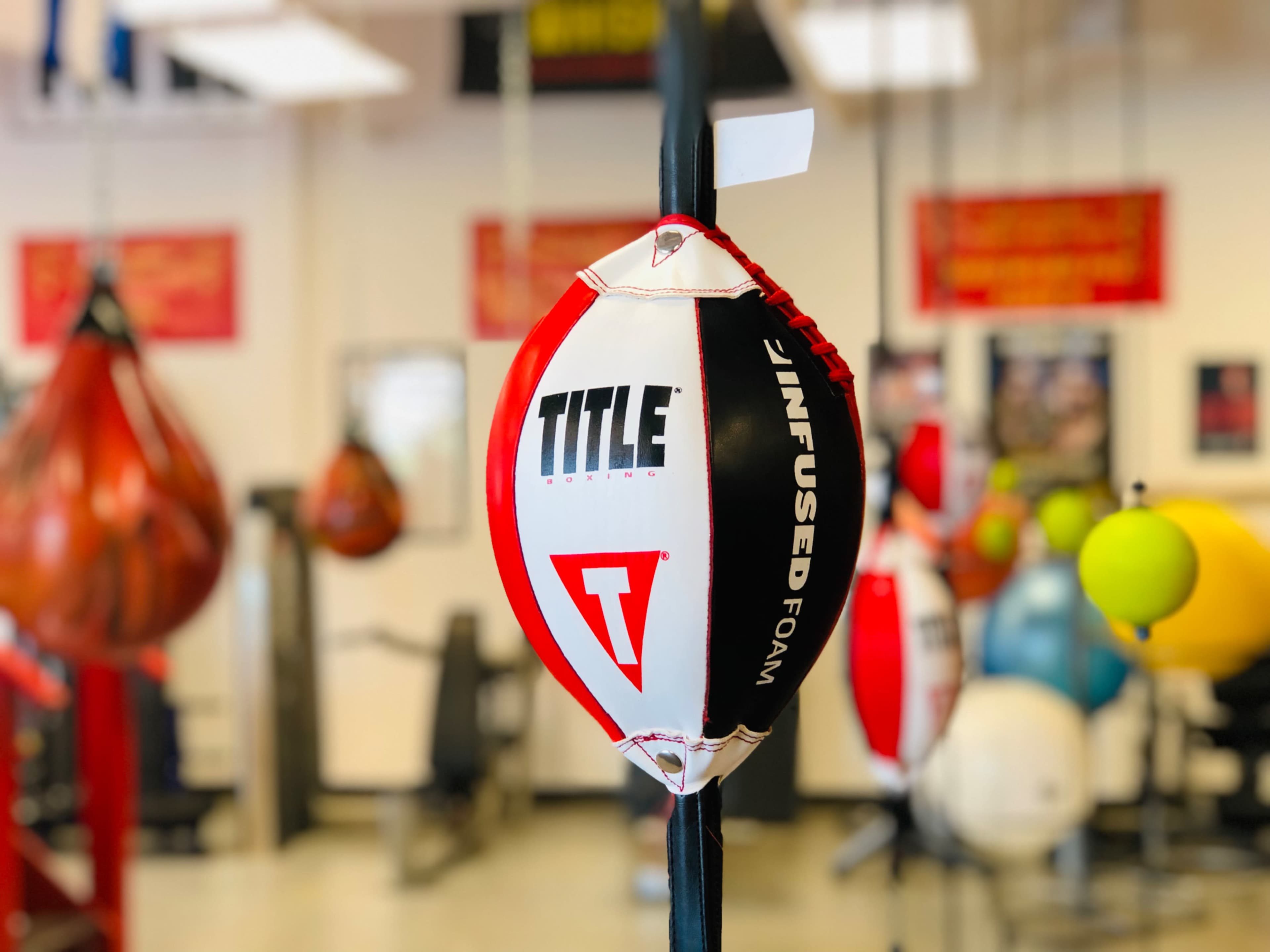 A red and white TITLE boxing speed bag hangs from a pole in a gym filled with various other exercise equipment.