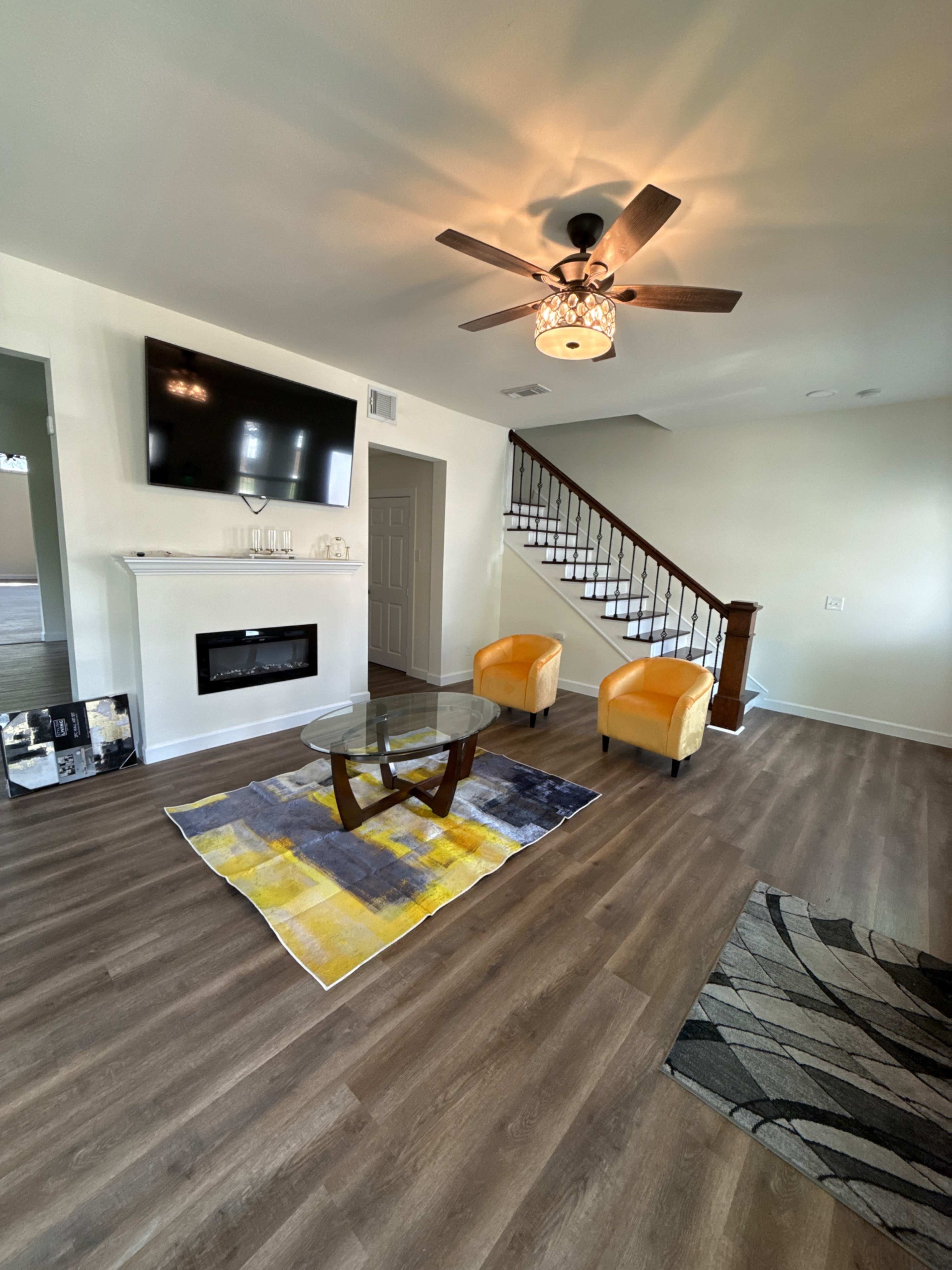 A living room features a ceiling fan, a mounted television, a fireplace, two yellow chairs, and a glass coffee table on an area rug, with a staircase in the background.
