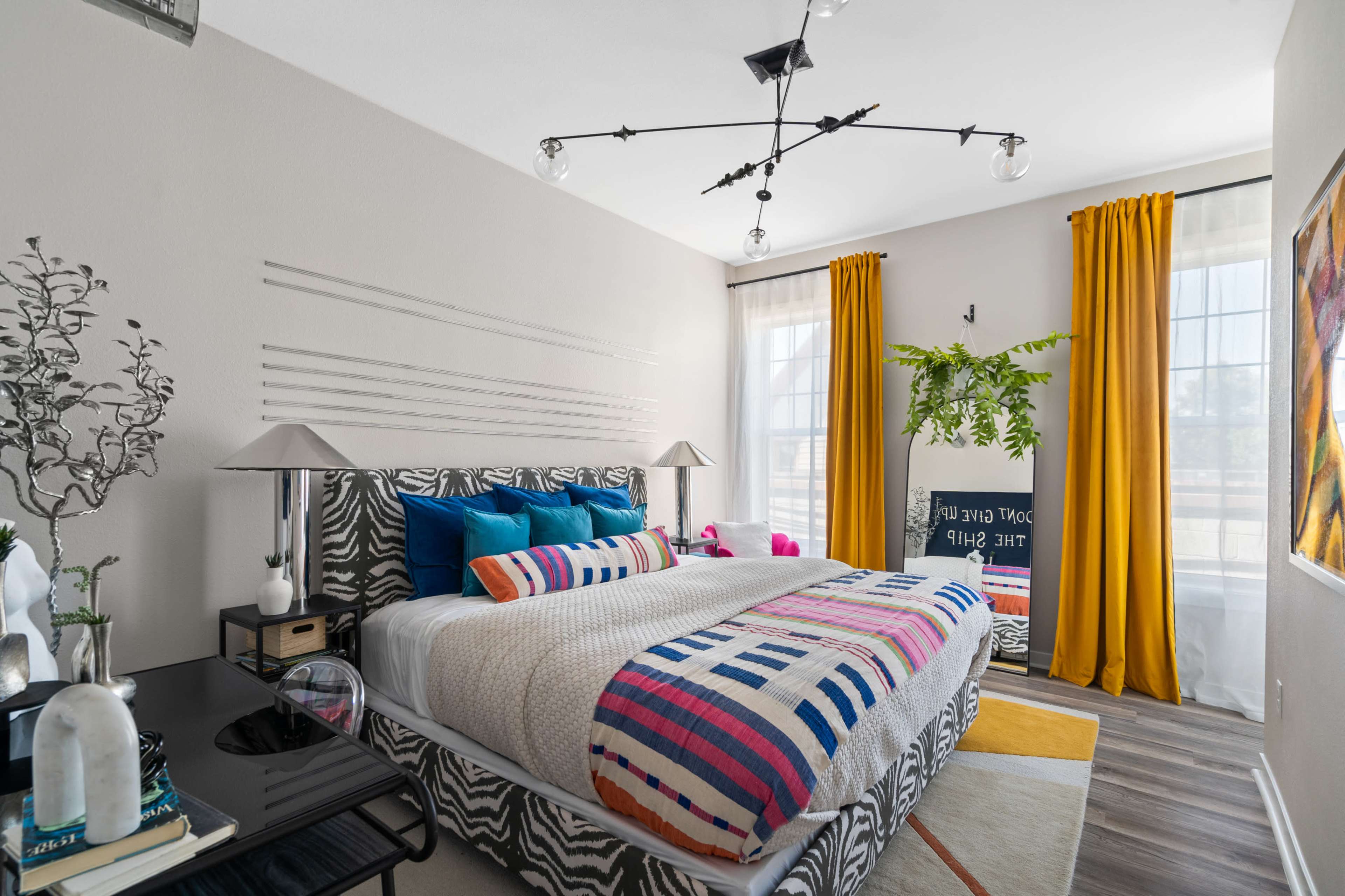 The image shows a modern bedroom with a patterned bedspread, colorful pillows, large yellow curtains, and a stylish ceiling light fixture.
