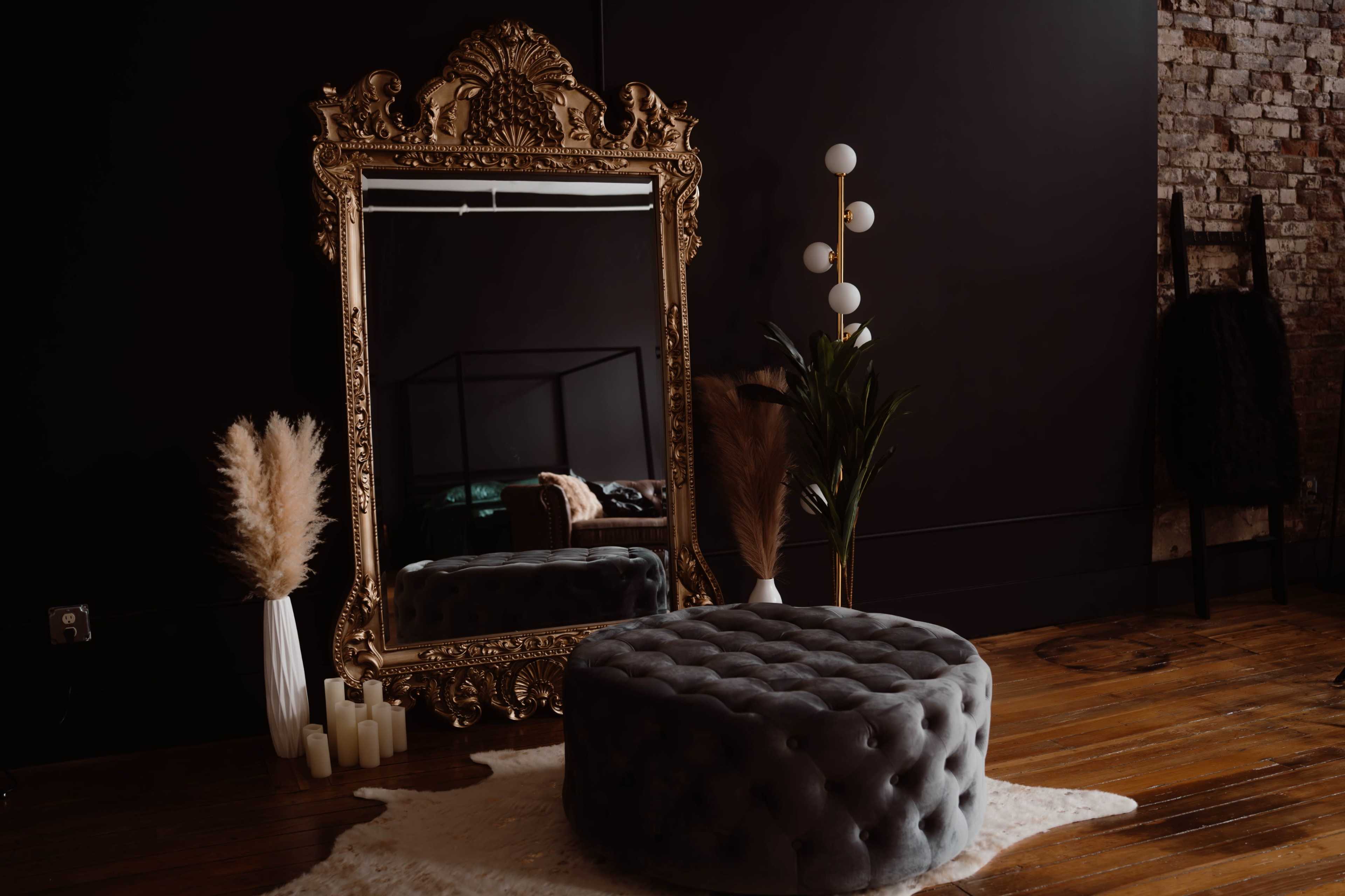 The scene features a large ornate mirror, a plush gray ottoman, a floor lamp with multiple globes, and decorative plants arranged on a wooden floor.