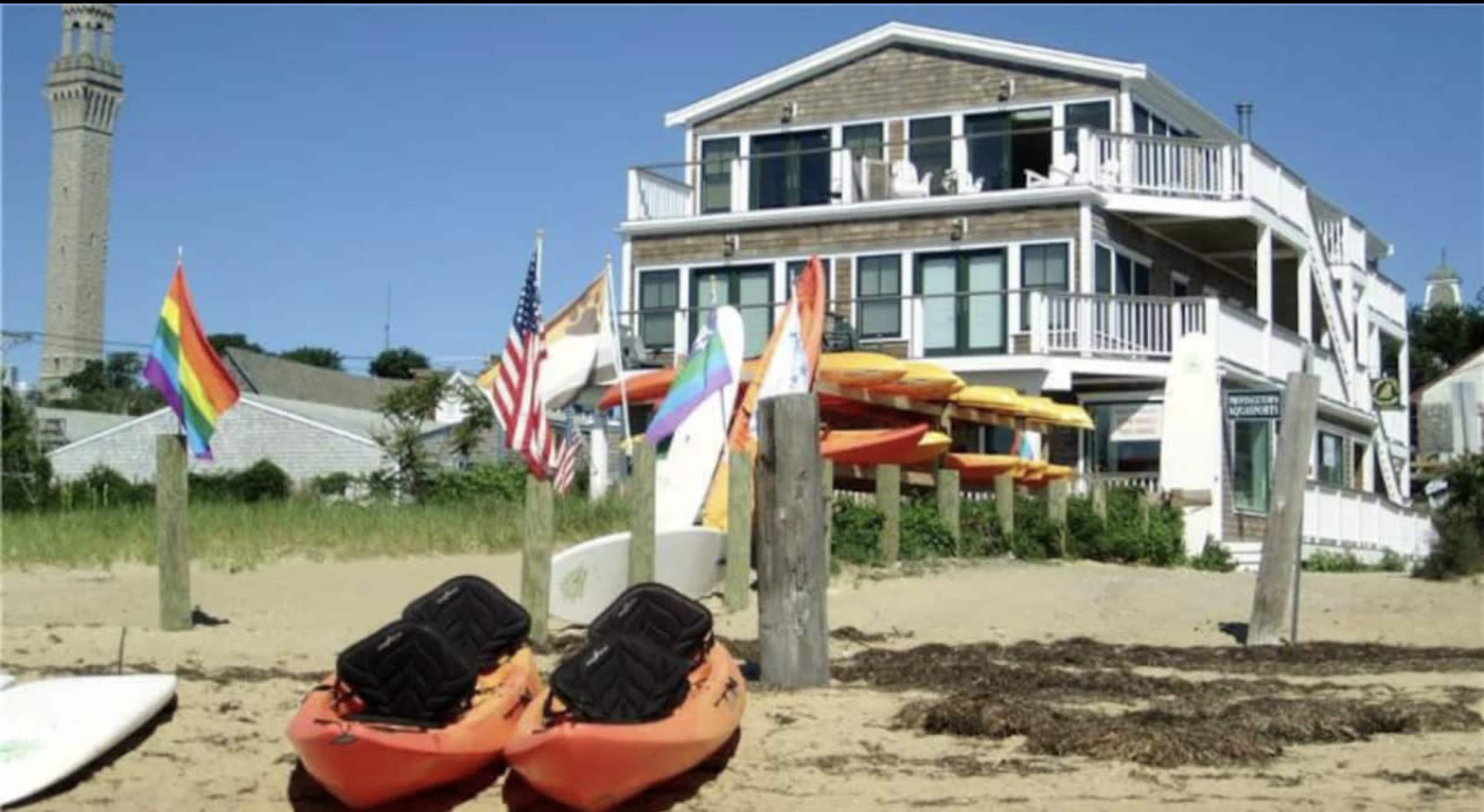 A beach scene with orange kayaks on the shore, colorful flags, and a multi-story building in the background.
