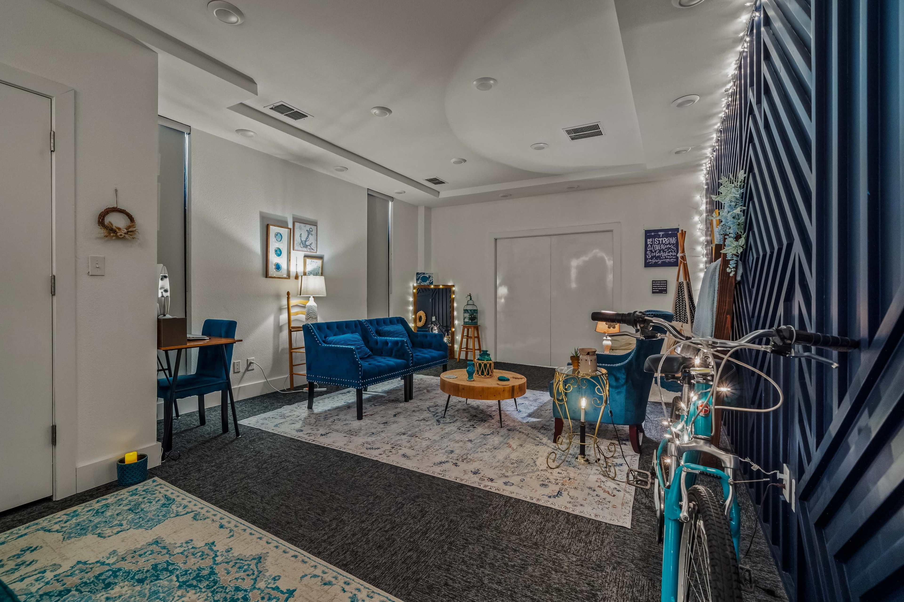 The image shows a modern, cozy lounge area with blue furniture, a wooden coffee table, a bicycle, and decorative lighting against a stylish backdrop.