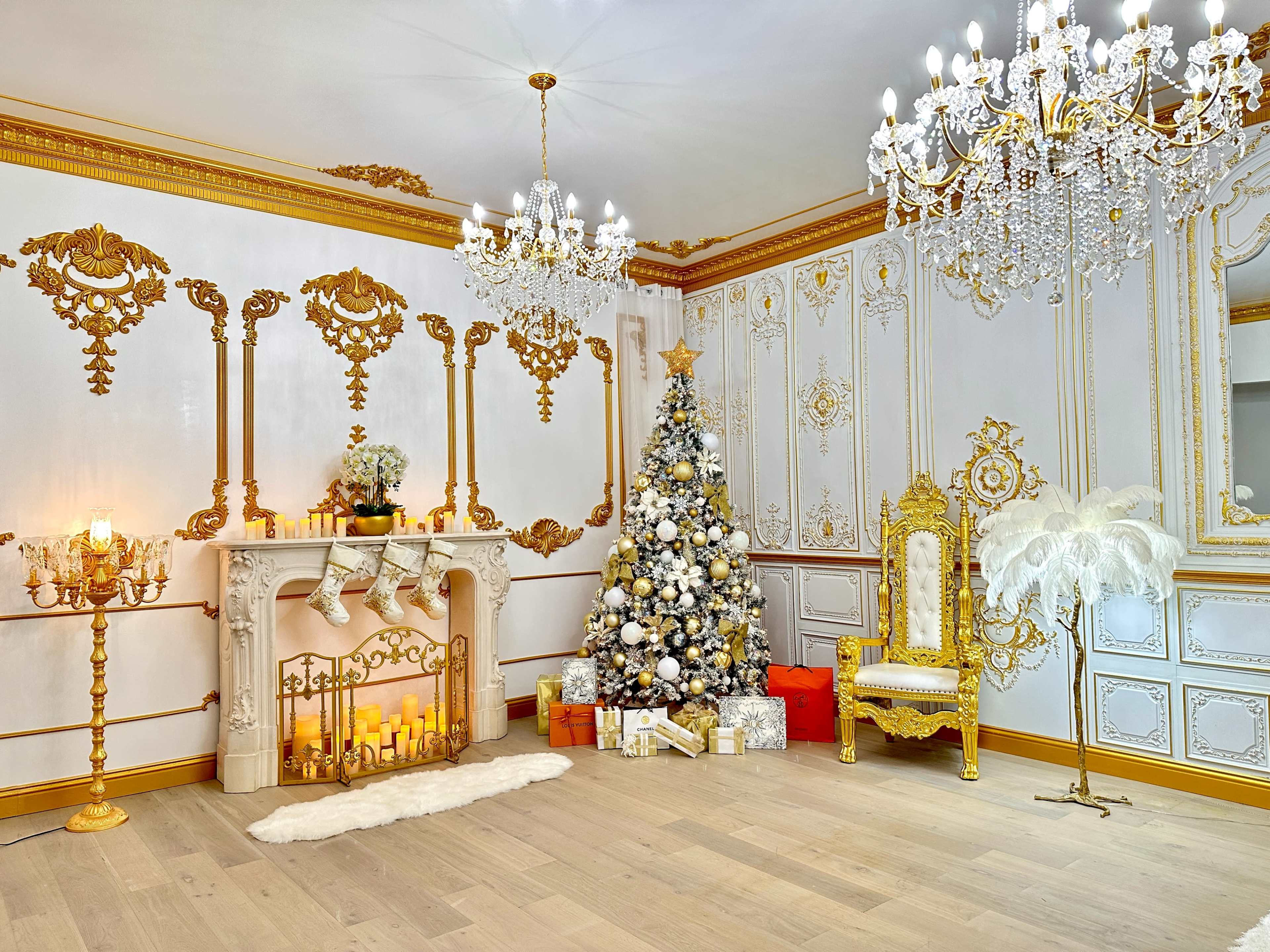 The image shows an elegantly decorated room with a gold-accented fireplace, a Christmas tree adorned with ornaments, and ornate chandeliers.