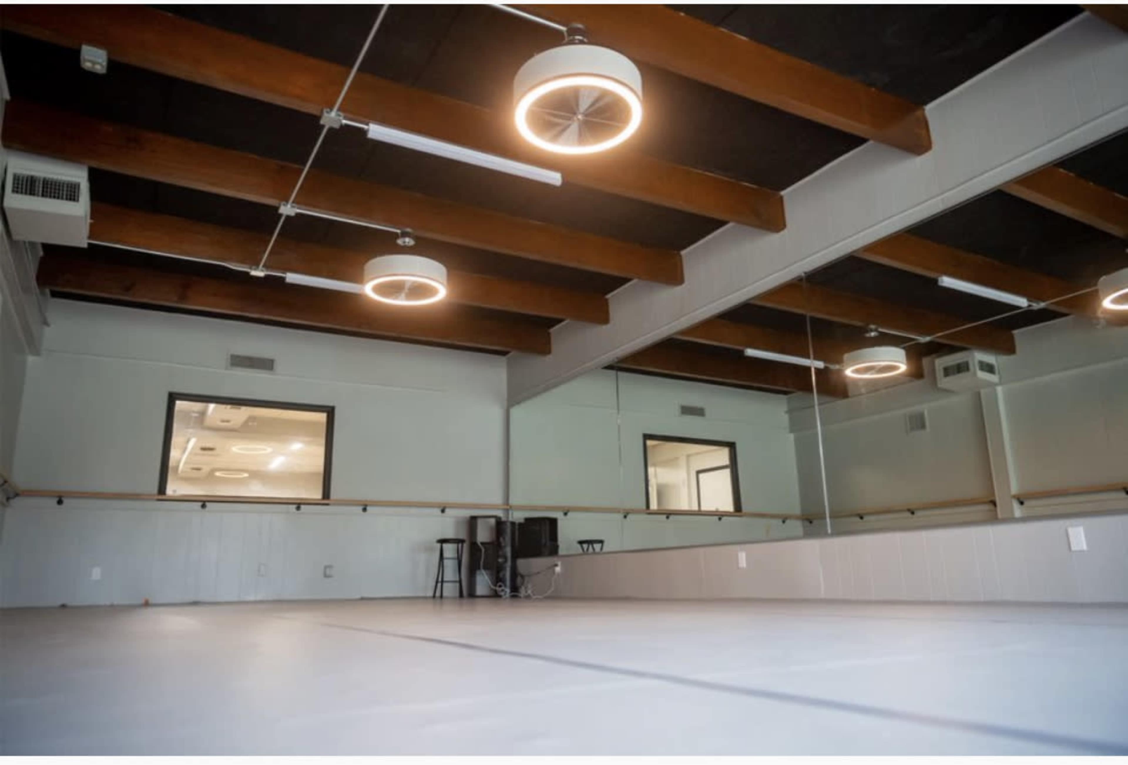The image shows an empty dance studio with wooden beams, circular ceiling lights, and a large mirror along one wall.