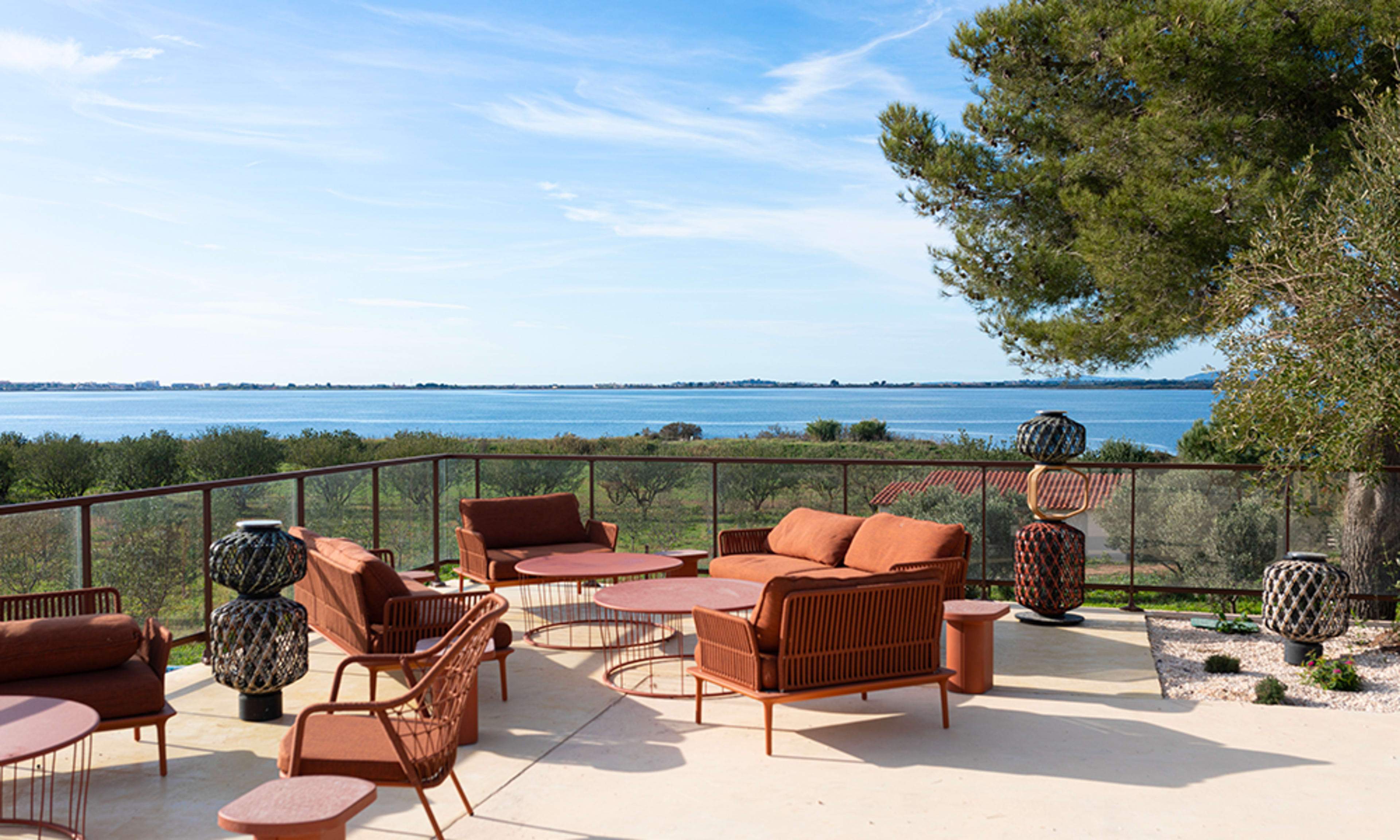 The image shows a terrace with orange furniture overlooking a body of water surrounded by greenery and trees.