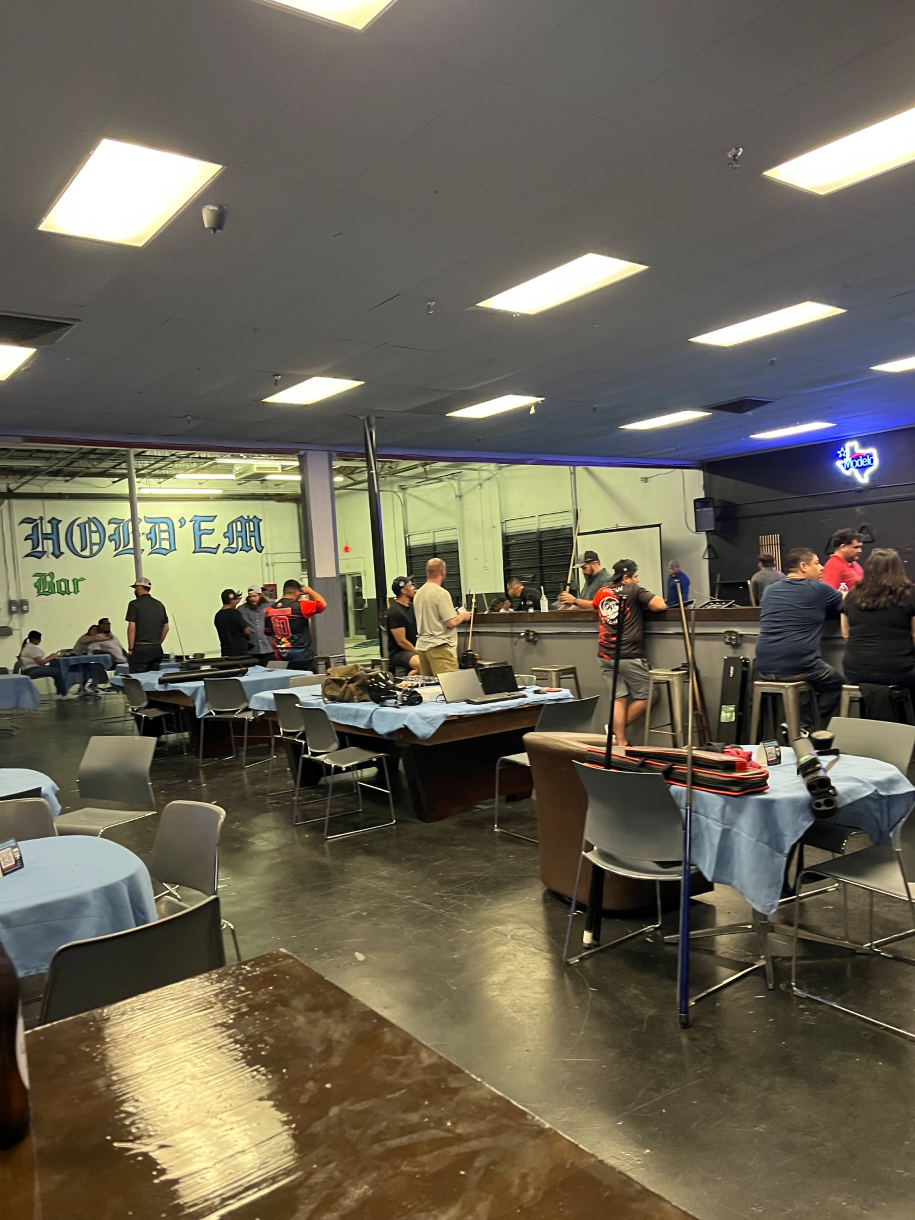 The image shows a spacious bar area with several tables set for patrons and a group of people gathered at the counter in a well-lit establishment.