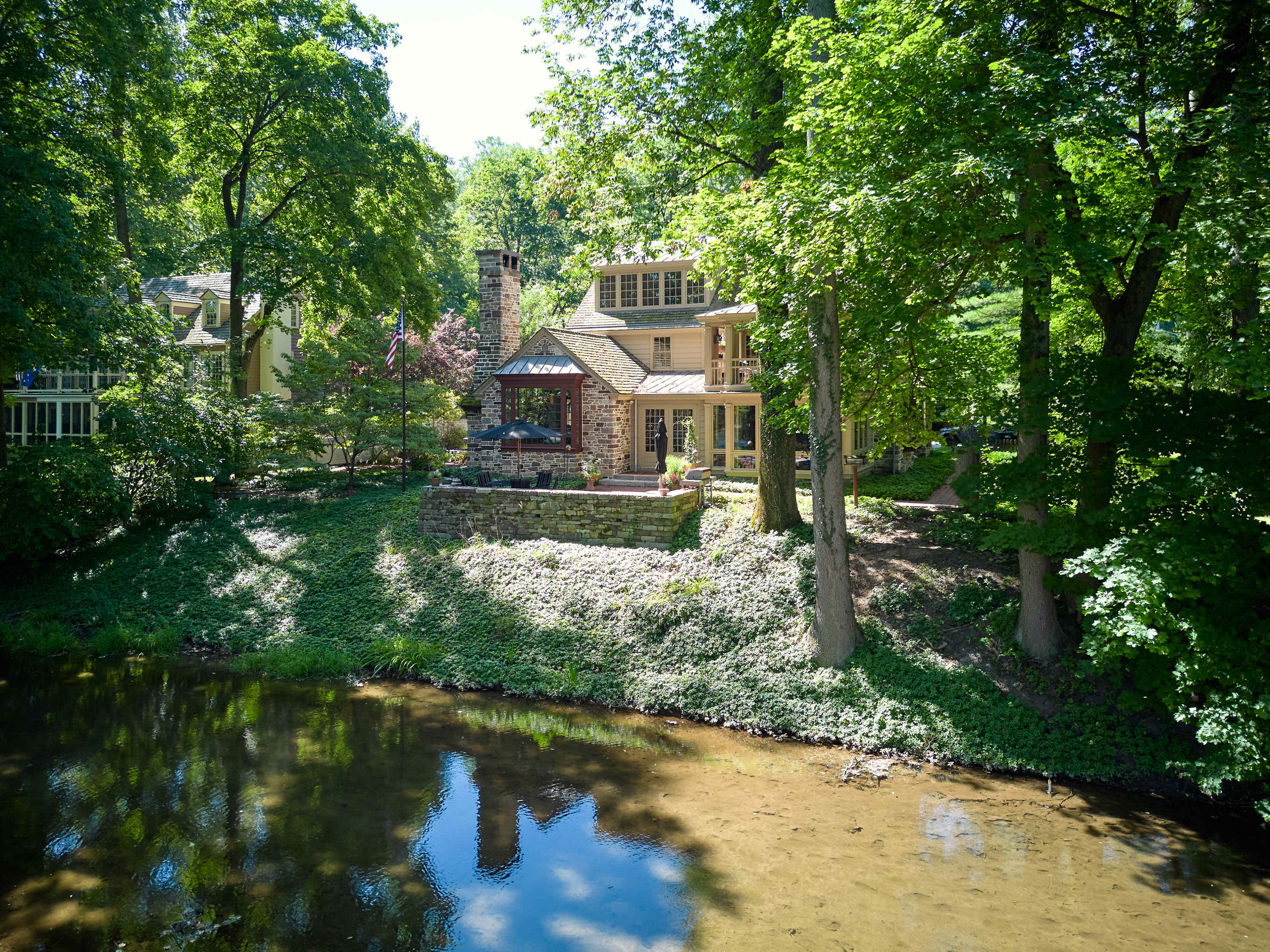 A two-story house made of stone and wood is situated near a clear stream, surrounded by lush greenery and trees.