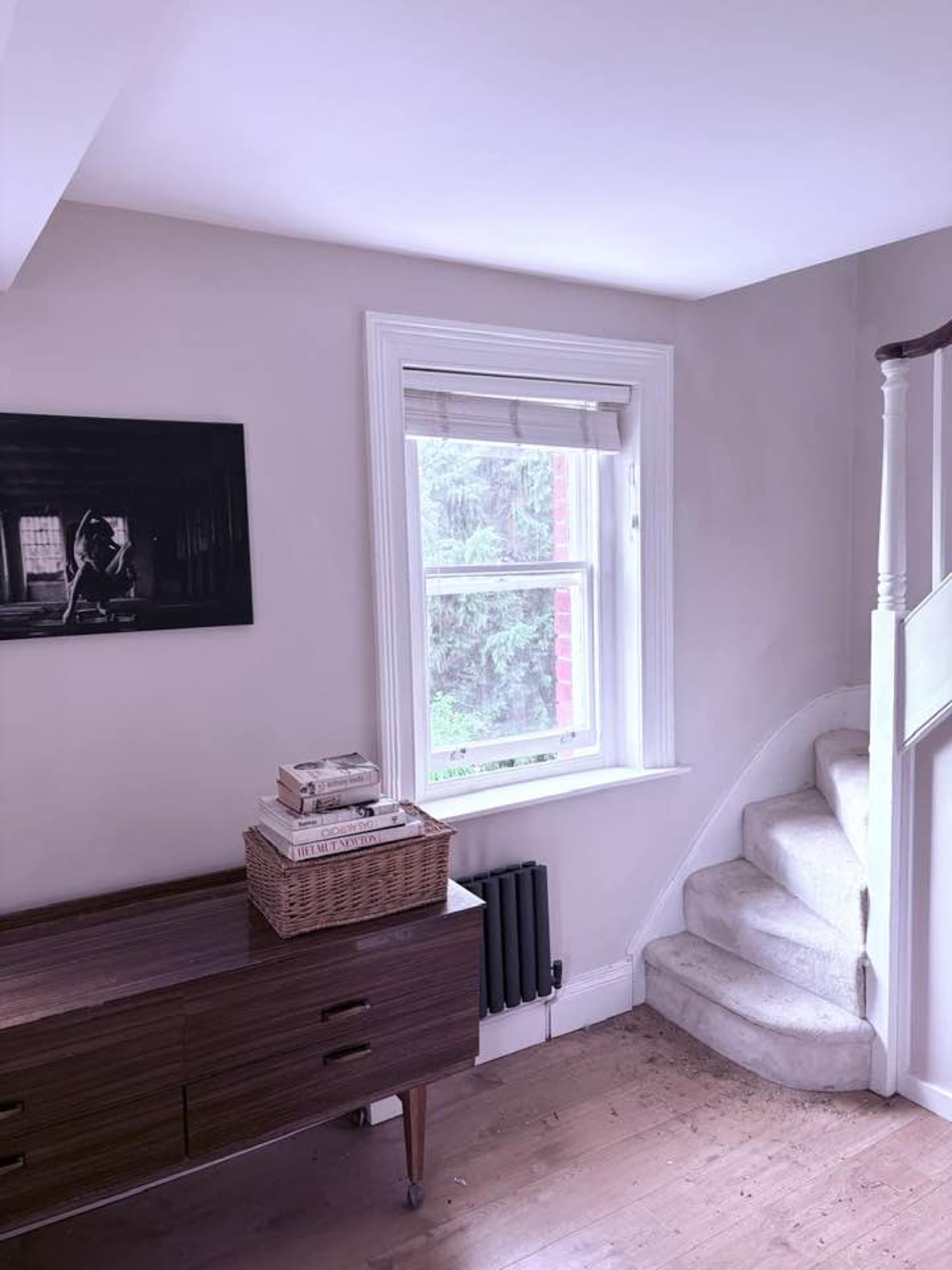 The image shows a corner of a room featuring a staircase, a window with blinds, a wooden cabinet with books and a basket, and a framed black-and-white photograph on the wall.
