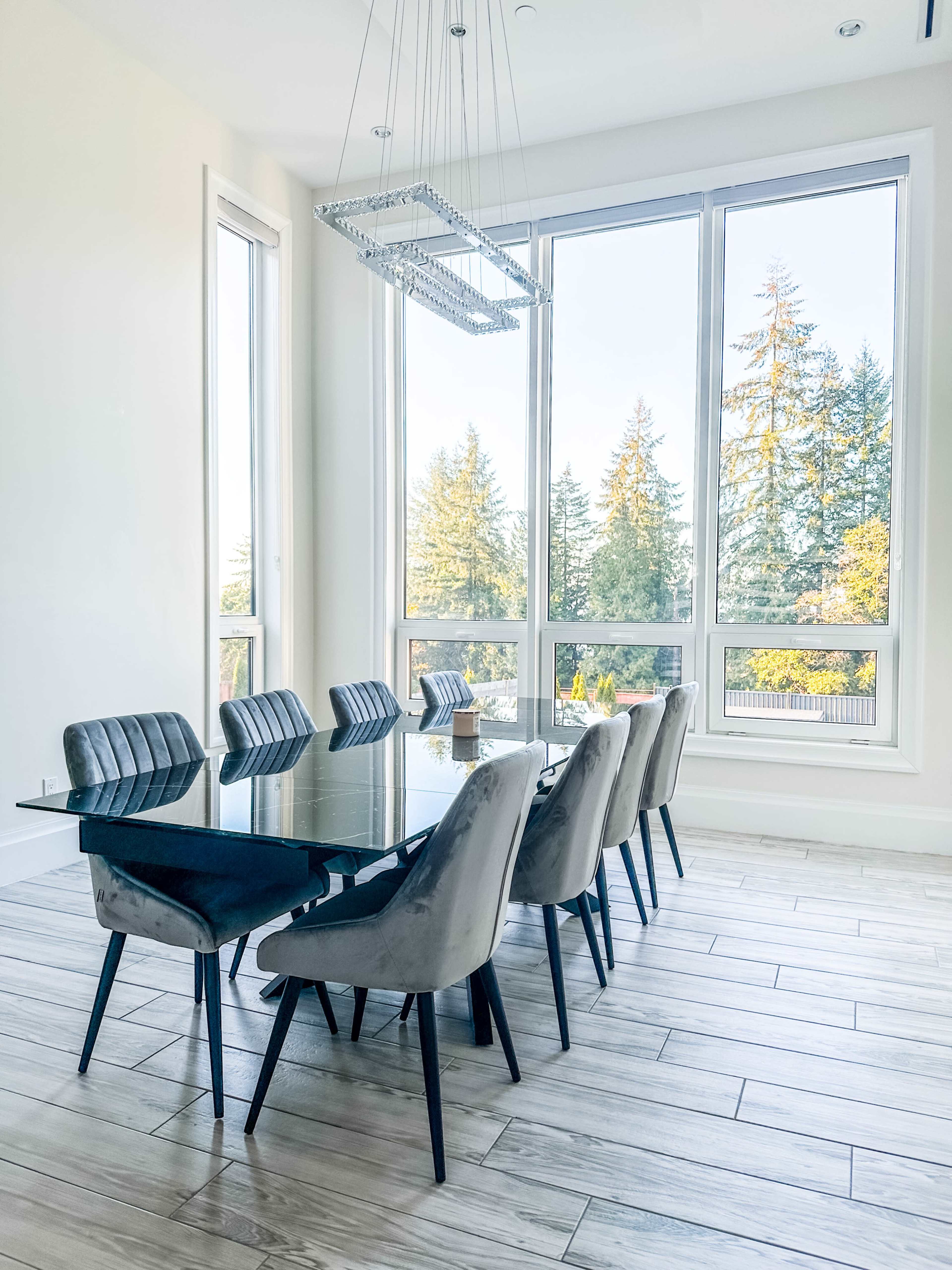 A modern dining room features a glass table surrounded by upholstered chairs, illuminated by a chandelier, with large windows showcasing trees outside.