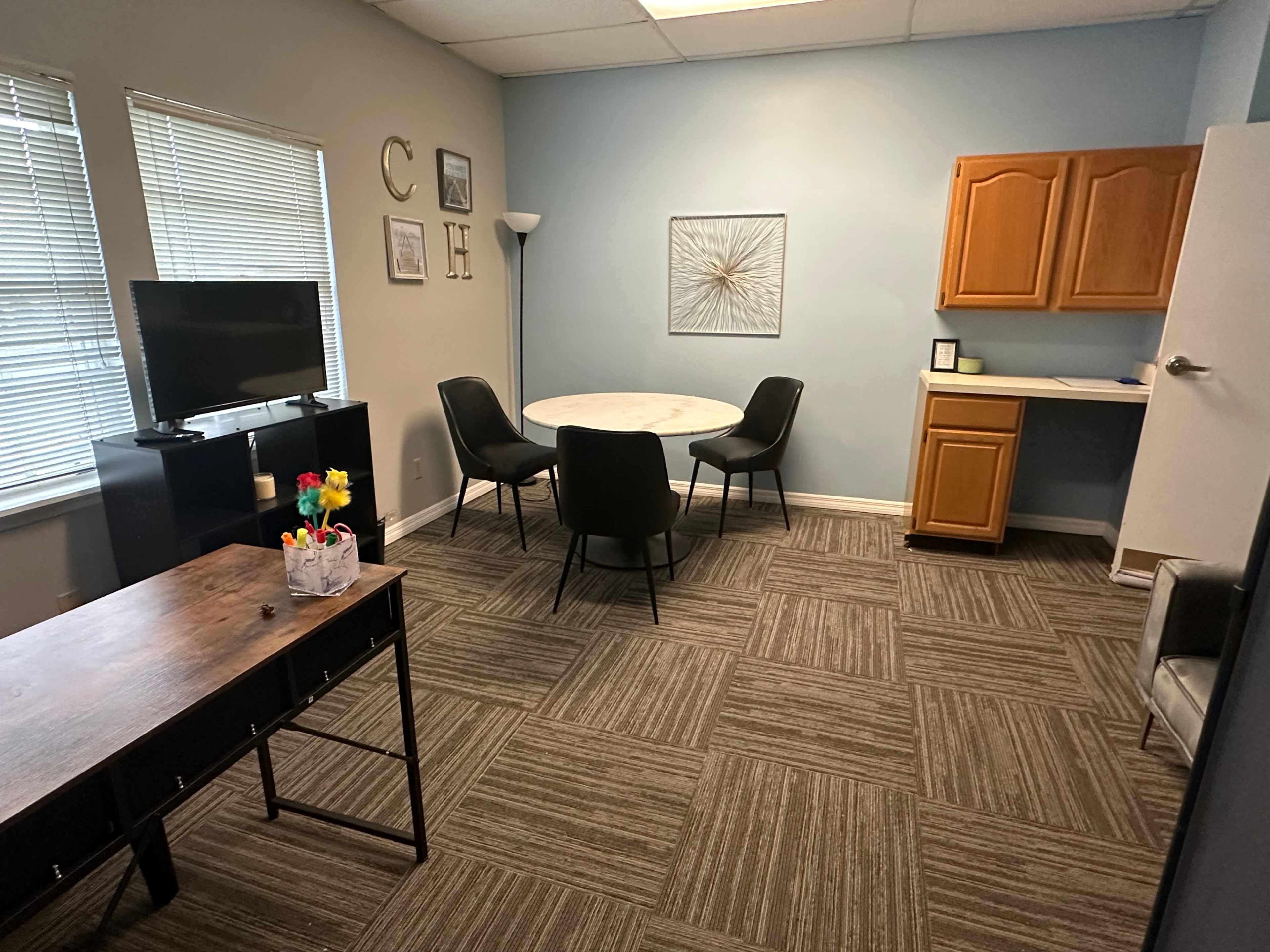 The room features a small dining area with a round table and four chairs, a television, a desk with office supplies, and a kitchenette with wooden cabinets.